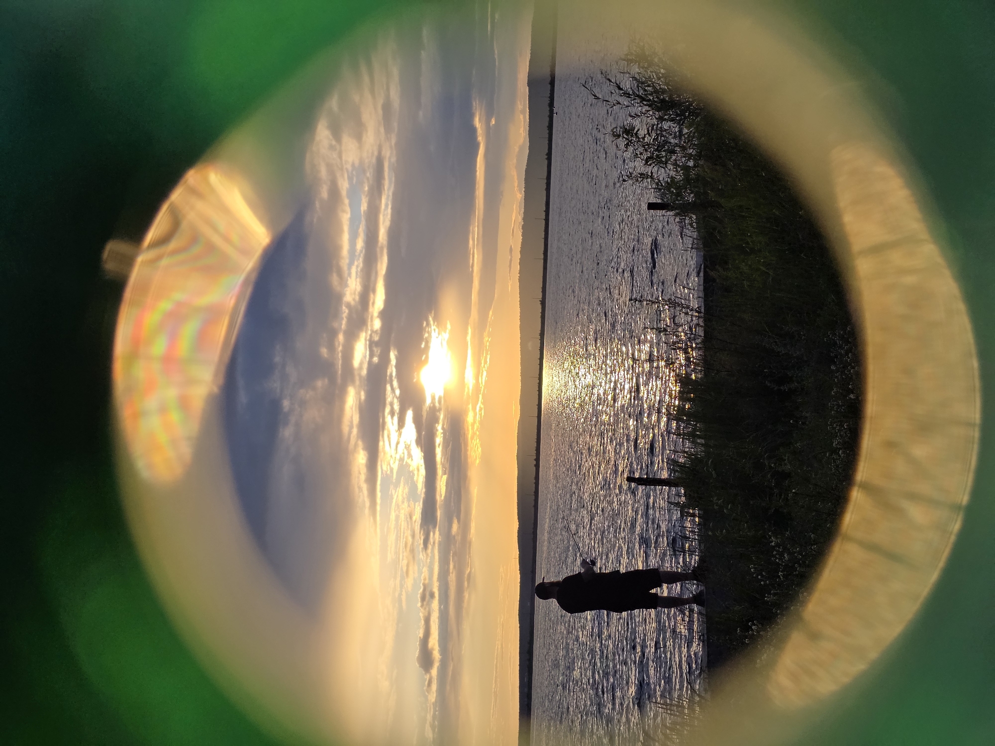 A person is fishing at the edge of a body of water during sunset. The sky is filled with dramatic clouds illuminated by the setting sun, creating a warm, golden glow. The water reflects the light and the sky, with ripples shimmering on its surface. The foreground shows a grassy bank with some reeds and a wooden post. The image is framed by a circular, out-of-focus lens effect, with green and yellow light flares on the edges. The overall mood is peaceful and serene, capturing a moment of quiet enjoyment in nature. The time of day is clearly sunset.