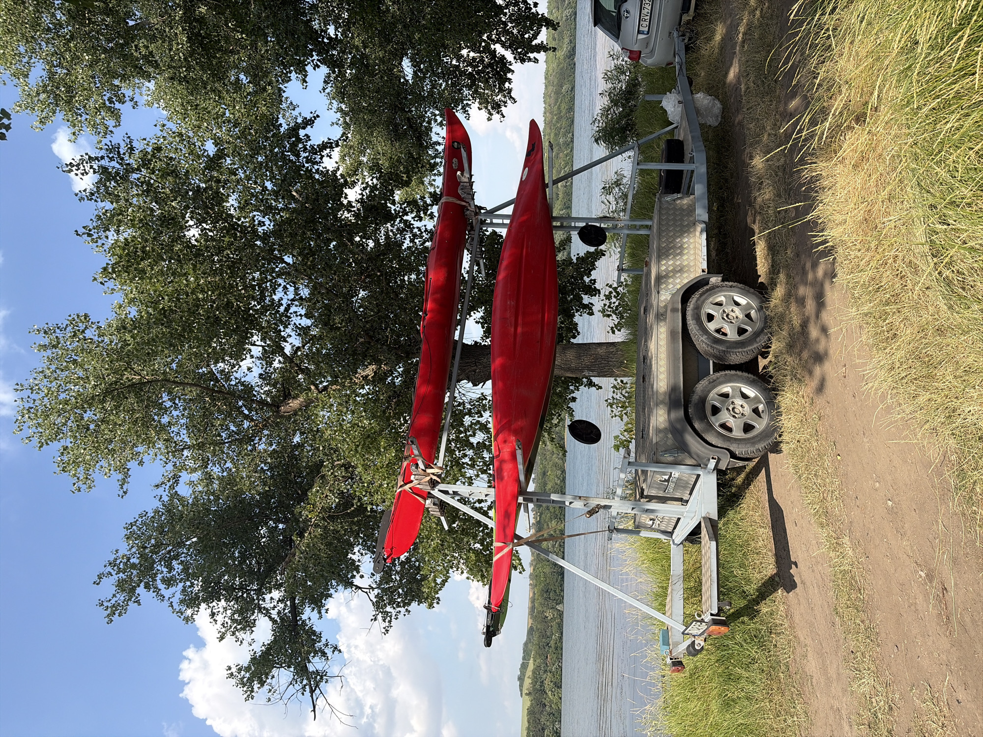 This is an eye-level, medium shot of two red kayaks on a trailer parked on a dirt path near a body of water. The kayaks are secured to a silver metal rack on top of a tandem-axle trailer. The trailer has two grey storage boxes on its deck. In the background, a large green tree with dense foliage looms over the scene, with a bright blue sky and scattered white clouds visible above. To the right of the trailer, a silver car is parked. The environment appears to be a natural setting with grass and a lake. The city of Oxentea, Moldova, is not visible in this image.