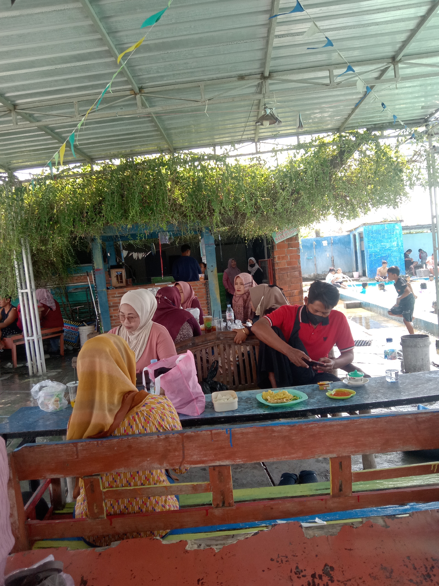 The image depicts an outdoor seating area, likely at a water park or recreational facility, in Kemamang, Indonesia. The foreground shows a weathered, multi-colored wooden bench. Behind it, a long, dark-colored table is set with plates of food, drinks, and cigarettes.
Several people are seated at the table, mostly facing away from the camera. One individual in a red polo shirt and black face mask is actively using a mobile phone. To the left, a person wearing a yellow hijab and patterned clothing is also seated at the table. A woman in a light-colored hijab and glasses is looking down at the table. Further to the left, other individuals are seated at separate tables, some under a covered structure.
The background reveals a bright blue swimming pool with several people, including children, swimming and playing. A building with blue walls and a tiled roof is visible adjacent to the pool. The area is shaded by a metal roof structure supported by poles, and draped with lush green hanging plants. Colorful triangular flags are strung across the ceiling of the covered area. The overall atmosphere suggests a casual, daytime gathering at a recreational venue.