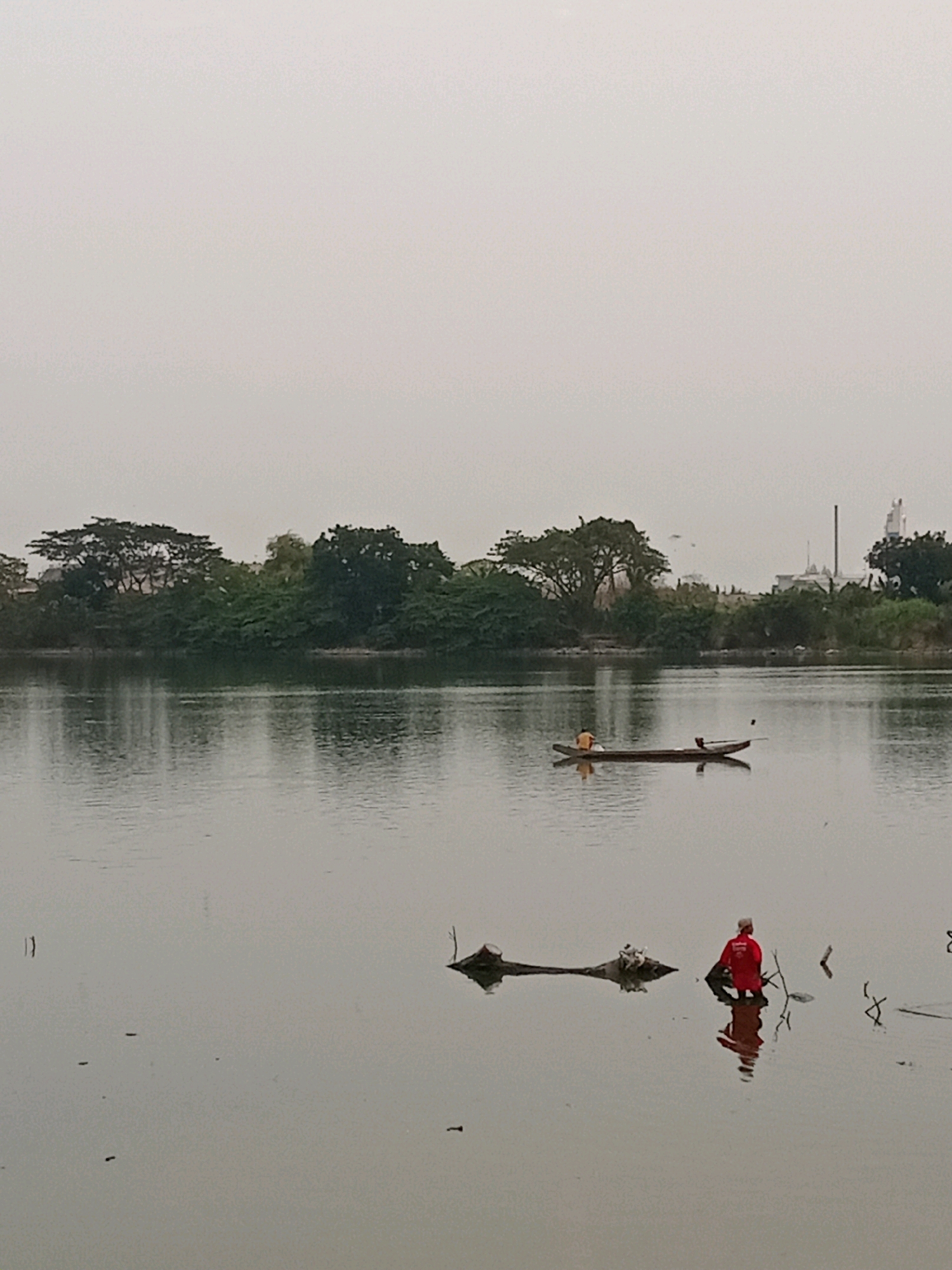 The image depicts a serene, overcast scene of a wide body of water, likely a river or a lake, adjacent to a lush green treeline. In the distance, silhouetted against the pale sky, are industrial structures, including a tall chimney, suggesting an urban or industrial area bordering this natural landscape in Karangturi, Indonesia.
In the foreground, a person wearing a bright red jacket and a white head covering stands in the shallow water amongst debris and driftwood. The person is facing away from the camera, appearing to interact with the debris. Further out on the water, a small wooden boat with two occupants, one in an orange shirt and another in a darker garment, glides across the surface. The boat is reflected in the calm water, as is the figure in red. The overall atmosphere is tranquil, with the soft light and still water creating a peaceful mood. The presence of both natural elements and industrial structures in the same frame highlights the coexistence of these two aspects in the environment.