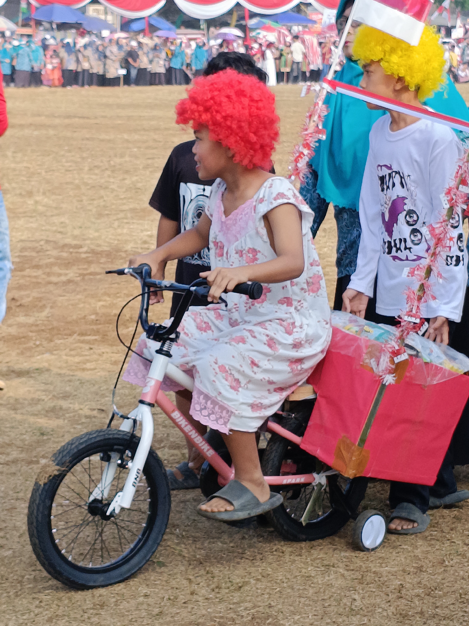 In Cibodas, Indonesia, a young child with a bright red afro wig sits on a pink and white bicycle, holding the handlebars. The child wears a white dress with pink floral patterns and grey sandals. A large red box adorned with decorative tassels is attached to the back of the bicycle. Another child, with a yellow afro wig, stands to the right, wearing a white shirt with a graphic print and holding a white and red Indonesian flag. Behind them, a crowd of people, many wearing blue or beige jackets, are gathered, with tents and umbrellas providing shade in the background. The ground is covered in dry grass and dirt.