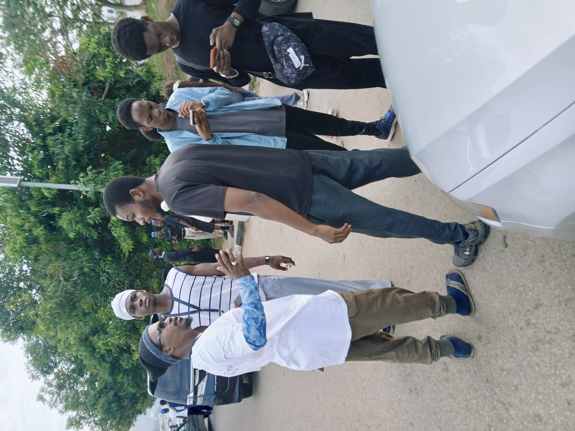 A group of people are standing and talking outdoors on a paved area, likely a street or parking lot, in the city of Unknown, Unknown. They are arranged somewhat closely together. One individual is wearing a black shirt, jeans, and dark sneakers. A second person is wearing a white T-shirt with blue sleeves and khaki pants. There is a Puma bag on the man dressed in black. A car is parked to the right of the frame. The location appears to have some vegetation in the background. Some individuals are holding and looking at phones.