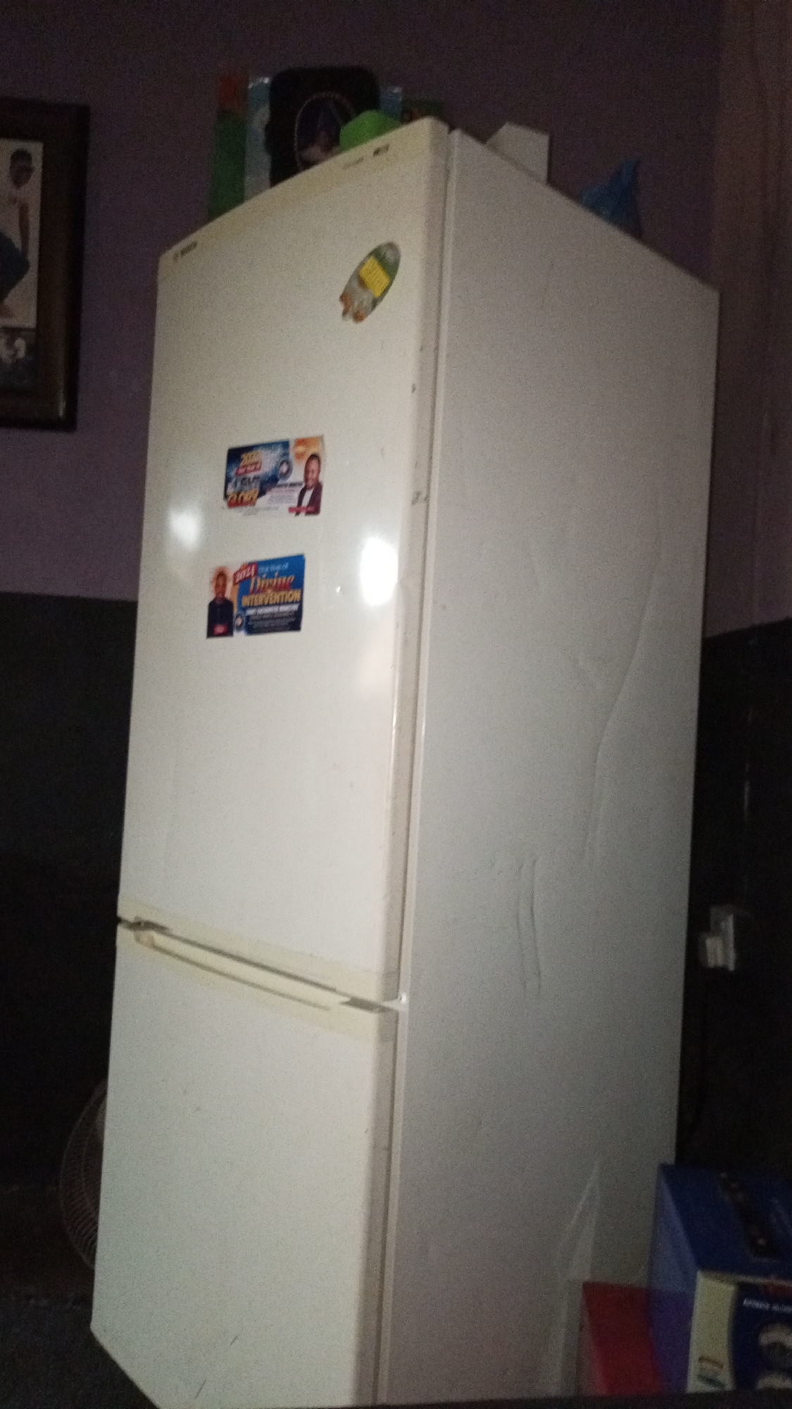The image depicts a white refrigerator with a top freezer compartment, positioned in a room. The fridge has several stickers and magnets on the front, including one that says "2023" and another labeled "Divine Intervention". Various items are stacked on top of the refrigerator. The surrounding environment appears to be indoors, with a framed picture visible on the wall to the left, and boxes are near the base of the refrigerator. An electrical outlet can be seen on the wall behind the fridge.