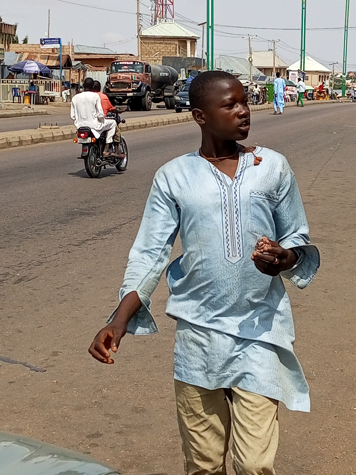A young boy walks on a paved street, facing right and looking slightly upward and to the right. He wears a light blue patterned tunic and tan trousers. In his right hand, he holds a small object. A thin brown necklace with two beads is around his neck. The street is lined with various buildings and shops, and there is traffic in the background, including a truck and a motorcycle with two passengers. The sky is clear and bright, suggesting a sunny day. The setting appears to be a busy urban or semi-urban environment.