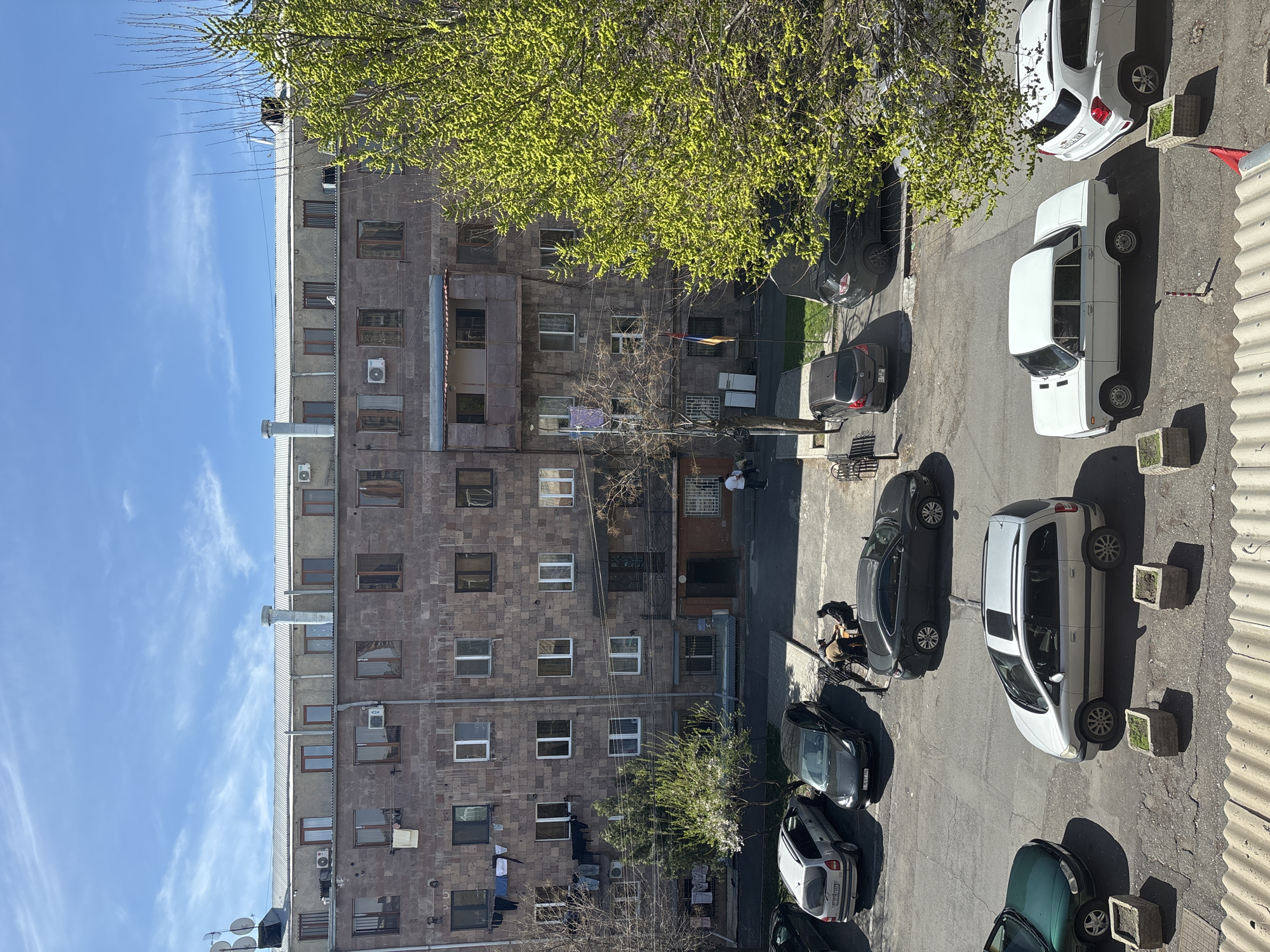 The image shows an exterior view of a multi-story residential building in Yerevan, Armenia. The building, constructed of a brown, rough-hewn stone-like material, features numerous windows and air conditioning units affixed to its exterior. Clotheslines extend from some windows, holding various articles of clothing. In the courtyard below, several parked cars occupy the space, including white, black, and green cars. Several people are present on the street. A tree with new spring foliage obscures part of the building. The sky is blue with sparse clouds. The image is taken from an elevated vantage point, looking down into the courtyard.