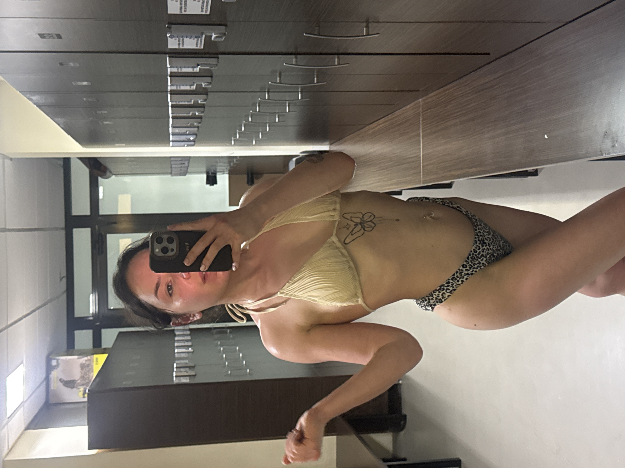 A woman is taking a selfie in a room with institutional furniture. The woman is wearing a light-colored bikini top and leopard-print bikini bottoms. She is holding a phone in her left hand, obscuring part of her face. There is a filing cabinet behind her. The room appears to be an office space with windows. The location and city cannot be determined.