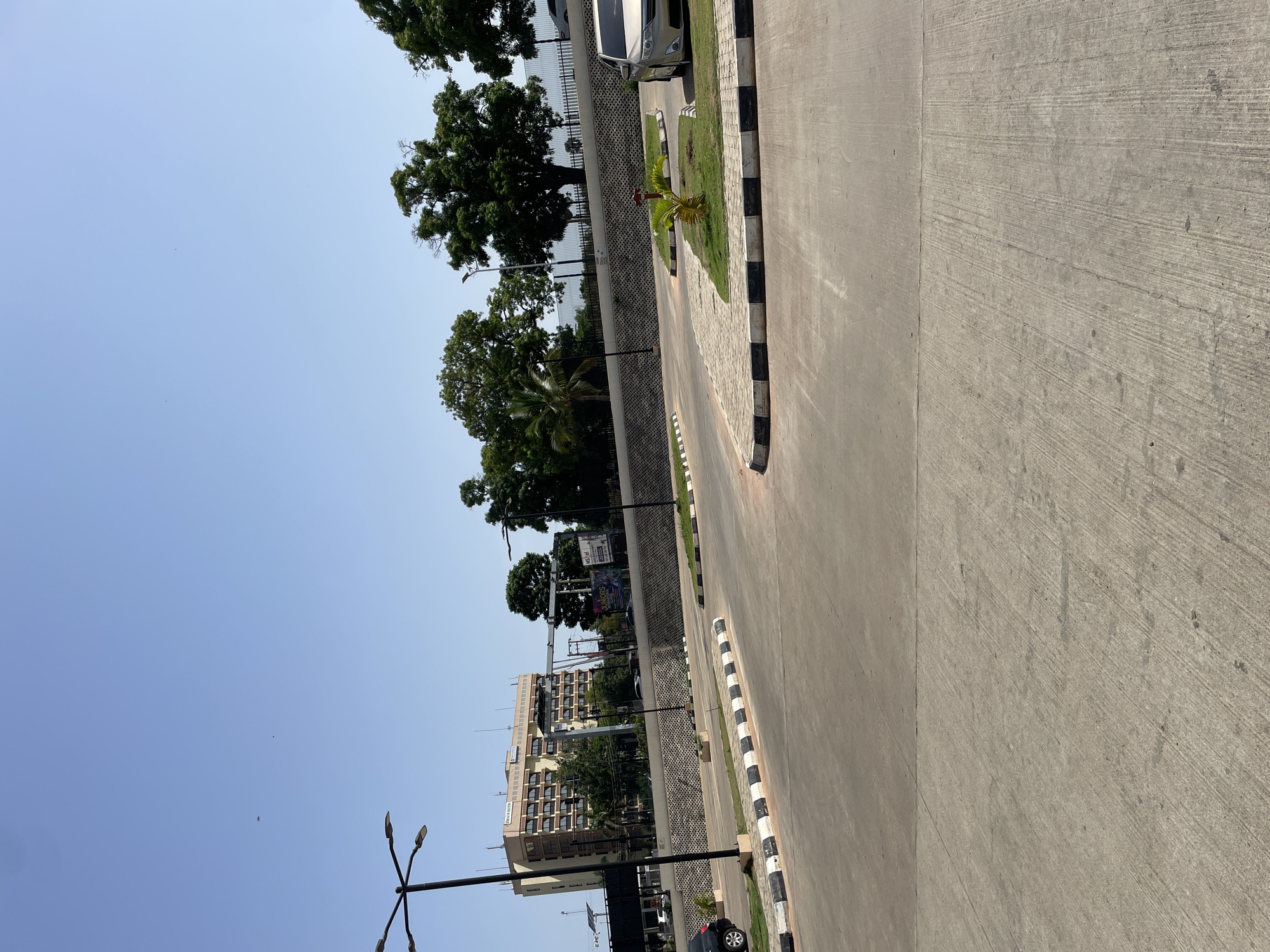 The image shows a street scene in Unguwar Rimi, Nigeria, on a clear day. In the foreground, a paved road with black and white striped curbs is visible, curving towards the right. Several lampposts line the road. In the middle ground, a low, patterned stone wall separates the road from a grassy area. Behind the wall, a modern, multi-story building with many windows stands, appearing to be a hotel or office complex. Trees of various sizes are scattered throughout the scene, some with dense foliage, others more sparse. A few cars are parked or driving on the road, including a silver sedan on the right and a black car partially visible on the left. Billboards with text and graphics are also present. The sky is a clear, bright blue.