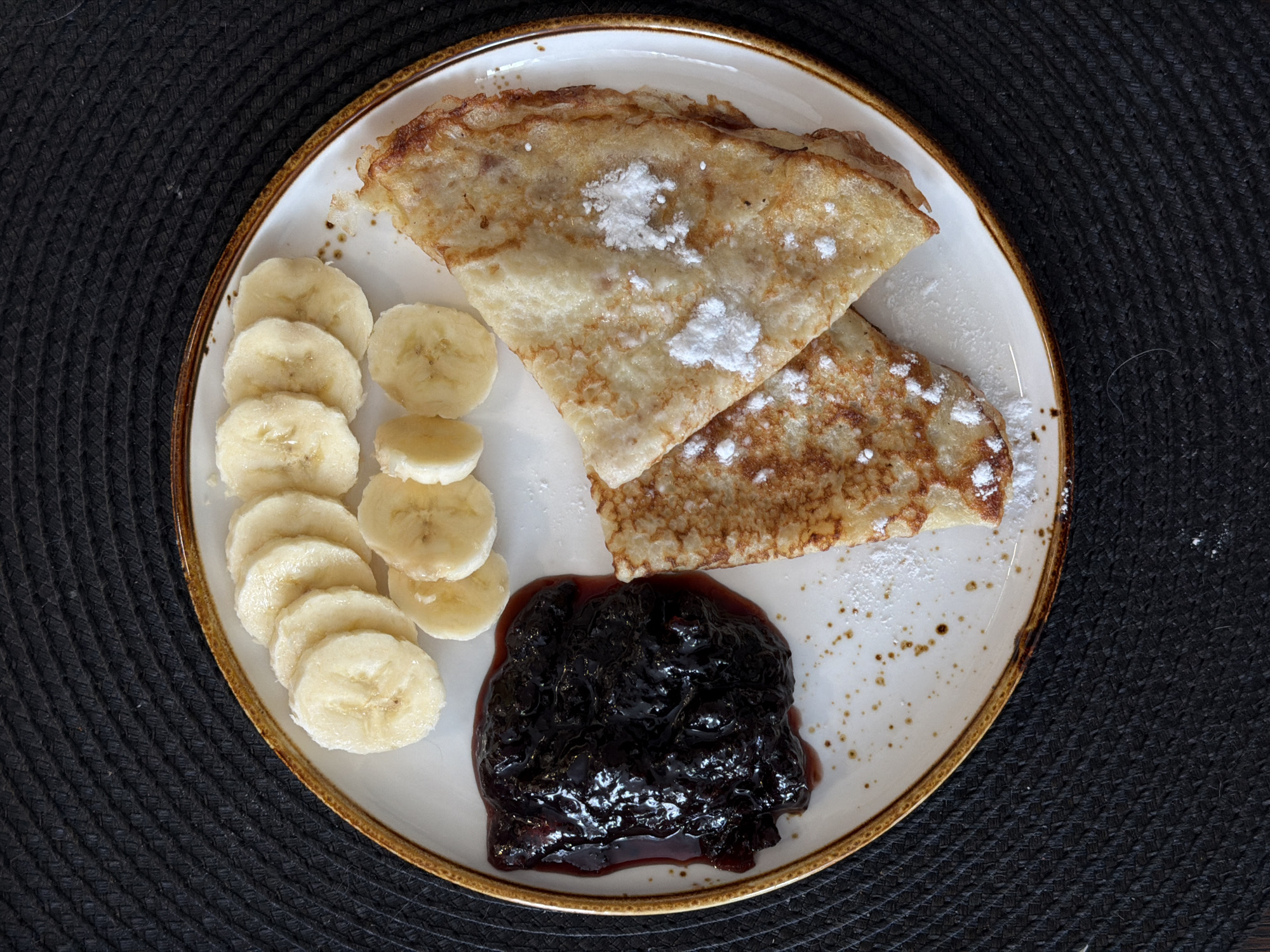 The image is an overhead shot of a white plate with a brown rim, containing a meal. The meal consists of two folded pancakes dusted with powdered sugar, a row of sliced bananas, and a serving of dark jam. The plate is set on a black, textured surface. The pancakes appear golden brown with slightly crisp edges. The banana slices are light yellow. The jam is a dark, glossy purple, likely made from berries. The background is uniformly black and textured. No people are visible in the image. There is no indication the photo was taken in Засечное, Russia.