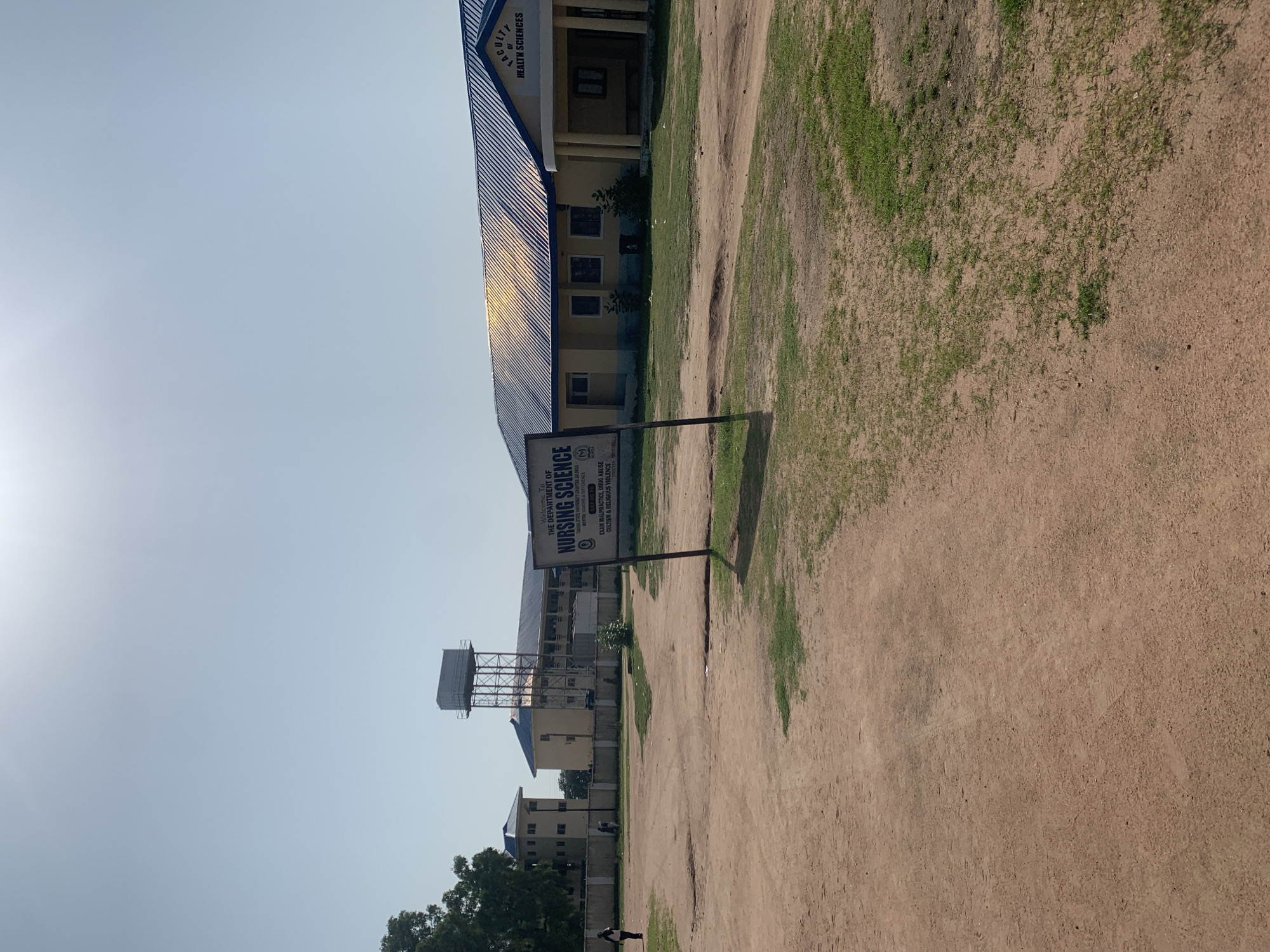 The image depicts the entrance to the Faculty of Health Sciences at Taraba State University in Jalingo, Nigeria. In the foreground, a dirt path with sparse green grass patches leads towards the buildings. A sign stands prominently, reading "WELCOME TO THE DEPARTMENT OF NURSING SCIENCE" and further listing "EXAM MALPRACTICE, DRUG ABUSE, CULTISM & RELIGIOUS VIOLENCE" as prohibited. To the right, a yellow building with a blue, corrugated metal roof is visible, with columns framing the entrance. Further back, a larger, multi-story building with a similar blue roof is seen, and a water tower with a tank on top stands tall. Two individuals are walking in the middle ground, near a wall that separates the campus from the surrounding area. The sky is clear and bright, suggesting a sunny day.