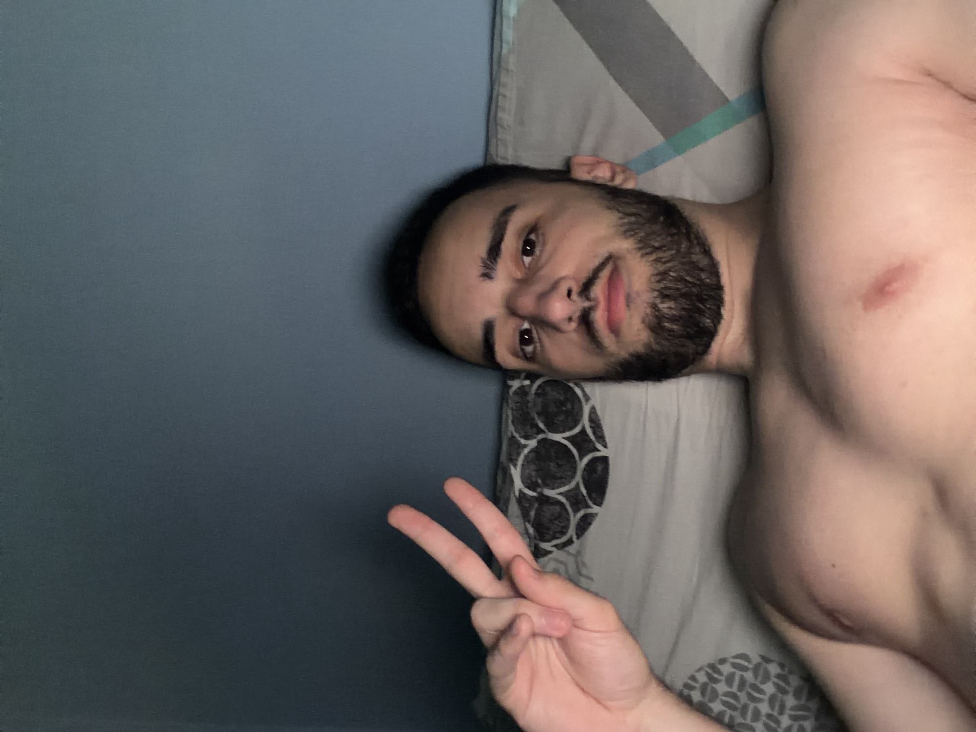 A man with a beard is lying on a bed, facing the camera and making a peace sign with his right hand. The background includes a grey wall and bedding. There is no information available to determine the location as Plovdiv, Bulgaria.