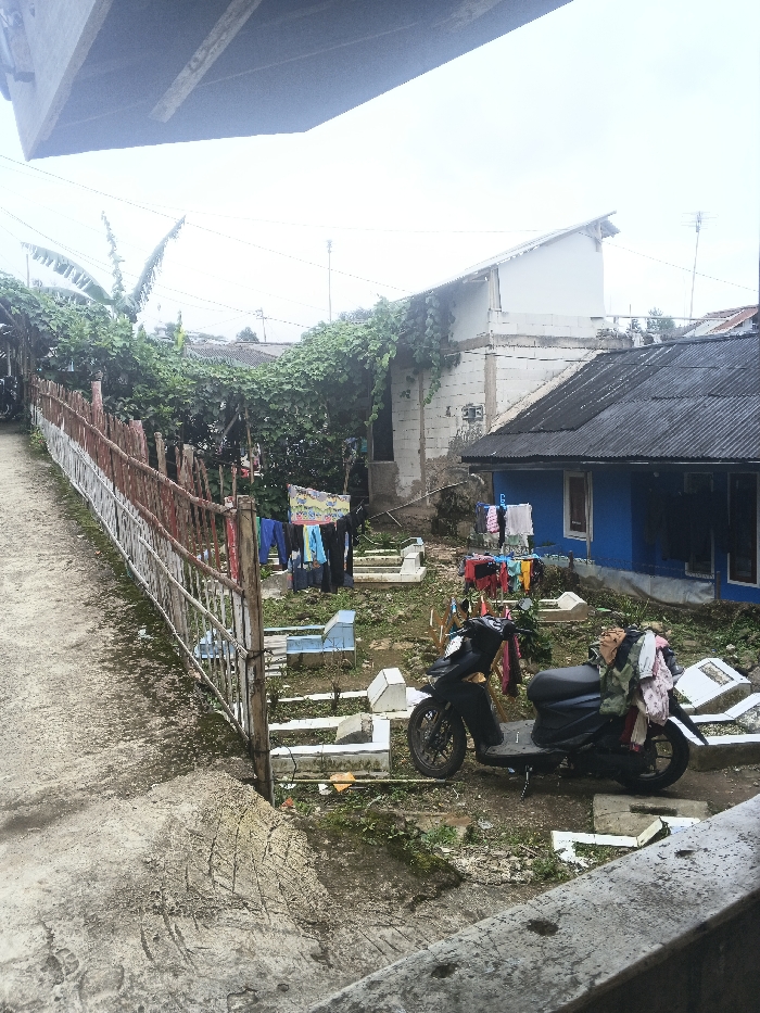 The image depicts an unkempt residential area bordering a burial ground in Bien Hoa, Vietnam, under an overcast sky. The upper frame is occupied by the dark, textured underside of an elevated concrete structure. A concrete path, displaying tire marks and patches of moss, slopes downwards on the left side, bordered by a fence composed of wooden posts and sections of red-and-white painted metal railings.
Behind this fence and path, an irregular plot of ground, sparsely covered with grass and weeds, contains multiple rectangular structures of white and light blue, identifiable as grave markers or small tombs. Two clotheslines are strung across this plot, holding various items of colorful laundry drying. A black scooter is parked facing left within this plot, resting on its kickstand, with colorful clothes draped over its seat and rear fender.
In the mid-ground, a single-story house with bright blue walls and a dark corrugated metal roof is visible on the right. To its left, a taller, unpainted concrete block building, partially overgrown with green vines, features a light-colored roof. Utility lines are visible above these structures in the background, set against a bright, featureless sky. No individuals are present in the scene.