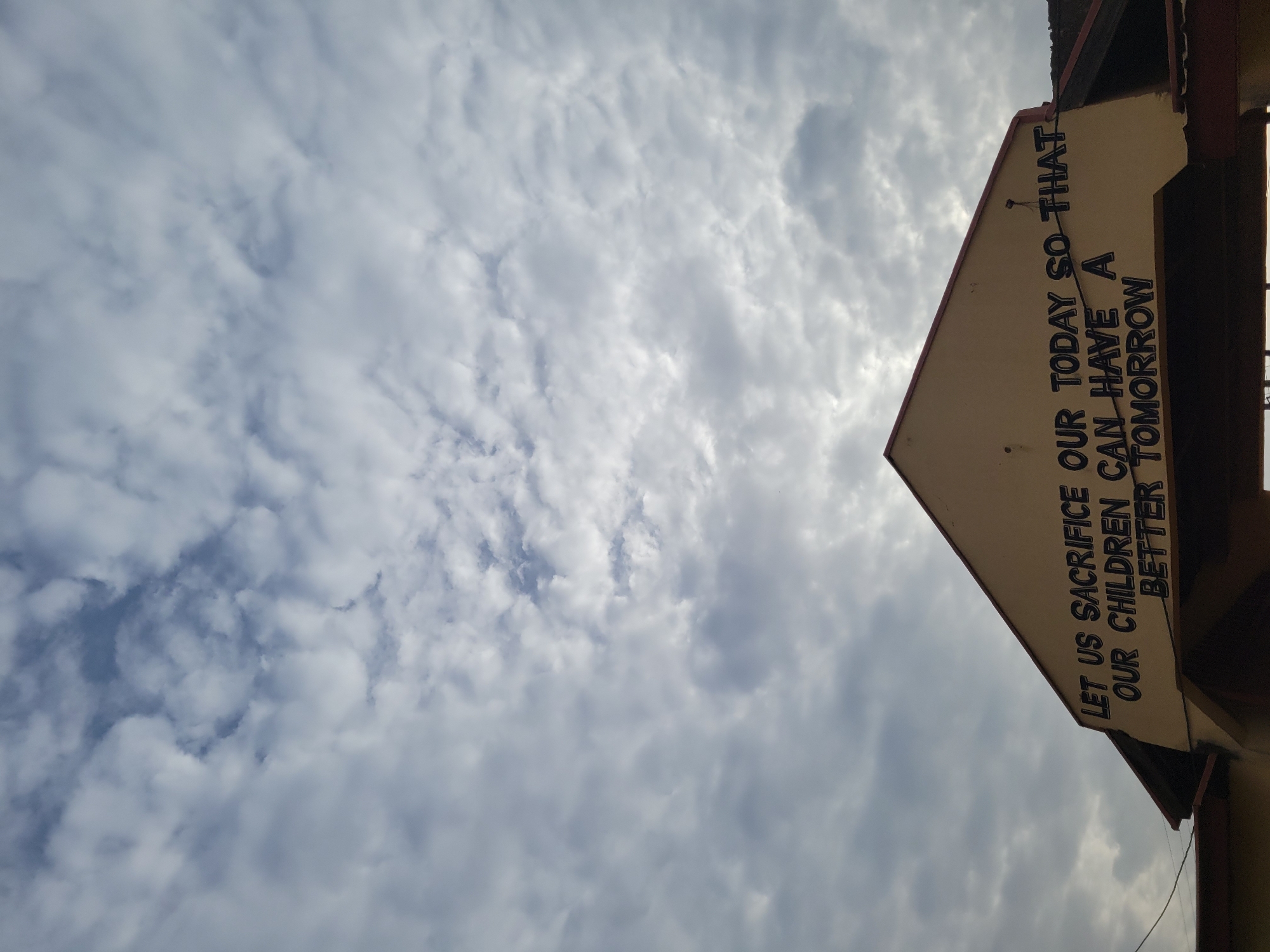 The image depicts a cloudy sky with a sign visible on the right. The sign reads, "LET US SACRIFICE OUR TODAY SO THAT OUR CHILDREN CAN HAVE A BETTER TOMORROW." The location is Gura Top, Nigeria.