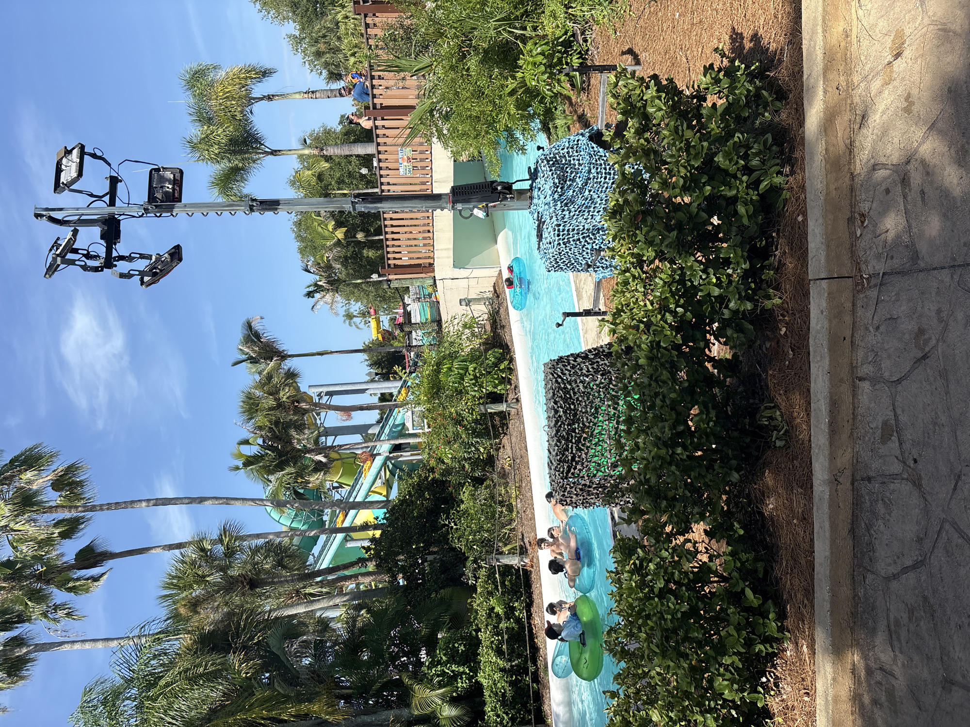 The image depicts a lazy river attraction at a water park, likely in Tampa, Florida, given the presence of palm trees and the style of the water slides in the background. Several individuals are floating in inflatable tubes on the turquoise water. One group of people, including children and adults, are clustered together in the foreground, with some wearing hats and swimwear. In the background, a vibrant green and yellow water slide is visible, hinting at the park's amenities. A tall, metallic pole with multiple lighting fixtures, possibly for night events, stands prominently in the middle ground. Dense green foliage lines the banks of the river, and a wooden fence separates the water park area from a pathway above. The scene is bathed in bright sunlight under a clear blue sky.