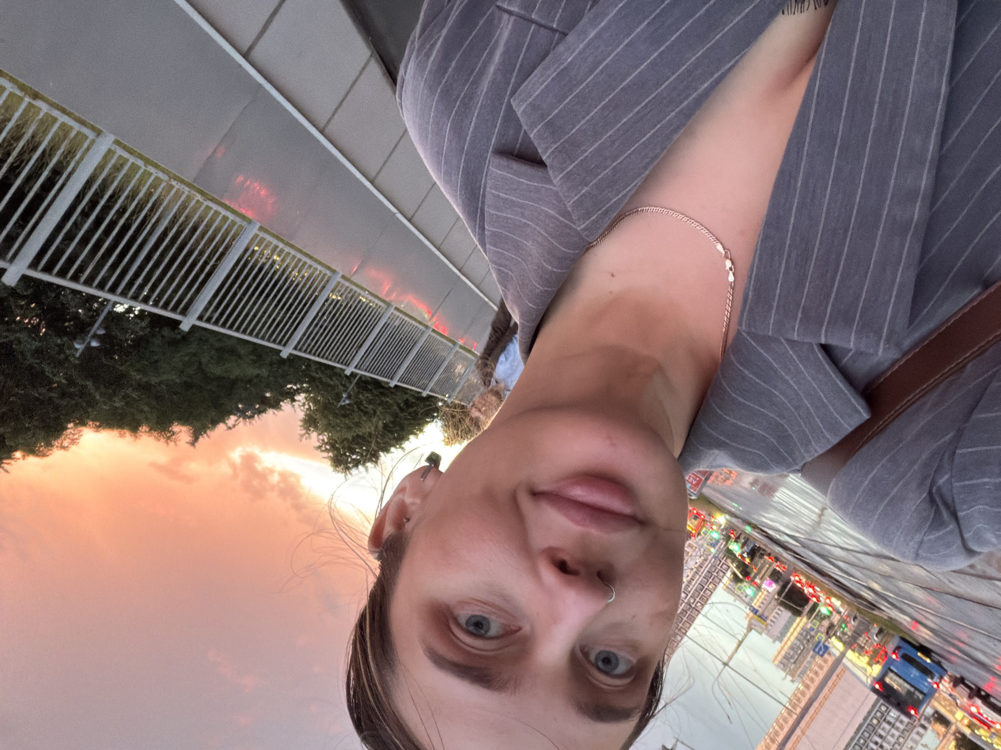 The media file is a selfie of a person outdoors at dusk. The sky is filled with colorful clouds, suggesting a sunset. The person is wearing a grey pinstripe blazer and a silver necklace. They have a nose ring and earrings. The background shows a white railing, trees, and a city street with lights and buildings, likely in Moscow, Russia. A blue bus is visible on the street. The overall mood appears to be casual and relaxed. The image is oriented sideways. There is no visible text.