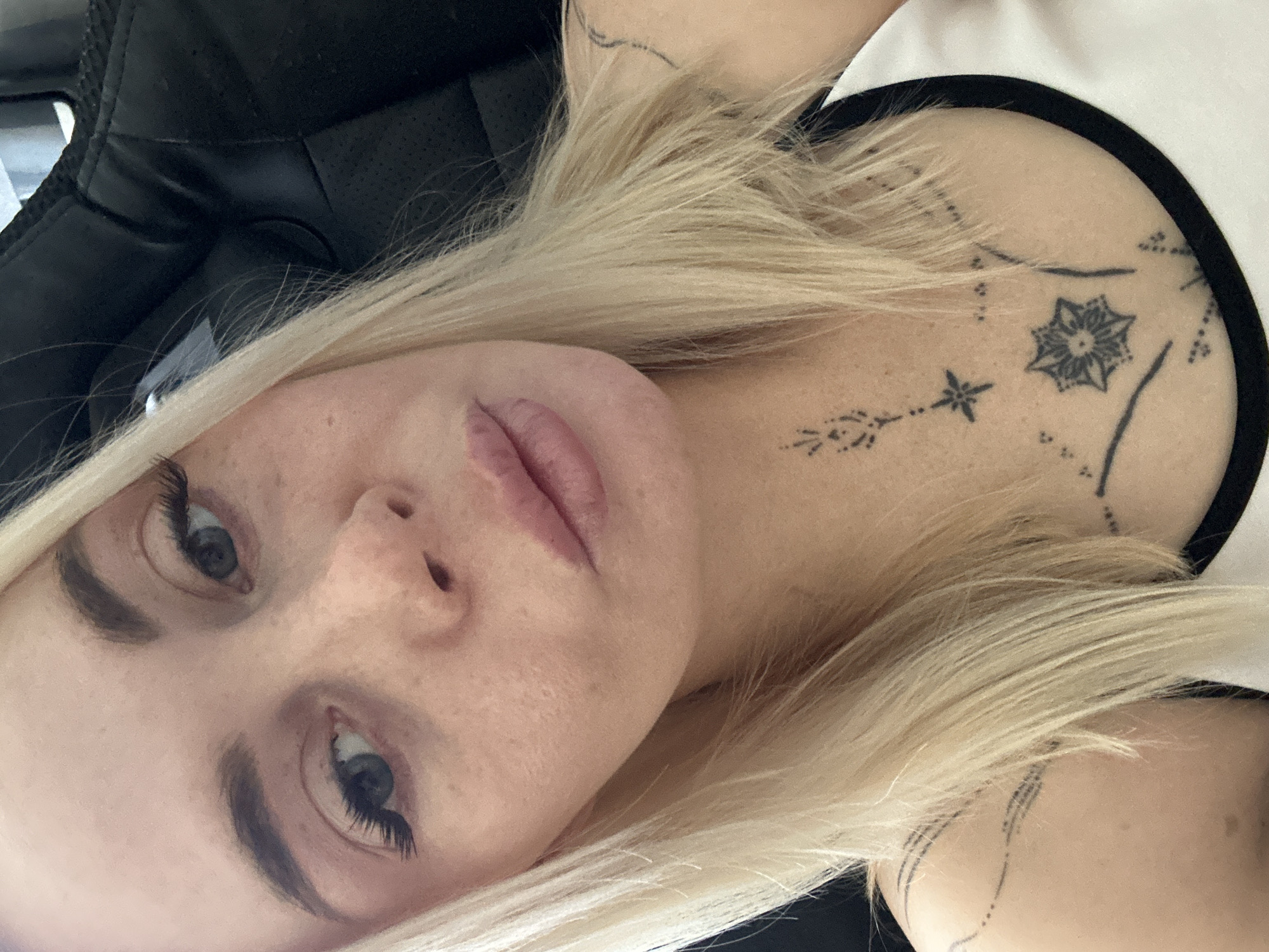 A close-up selfie shows a fair-skinned woman with blonde hair and blue eyes. She has light makeup, including mascara and lip gloss. She is wearing a white tank top with a black trim. She has several tattoos, including a starburst design on her chest and abstract lines on her shoulders and collarbone. She is seated in a black chair, the background is out of focus.