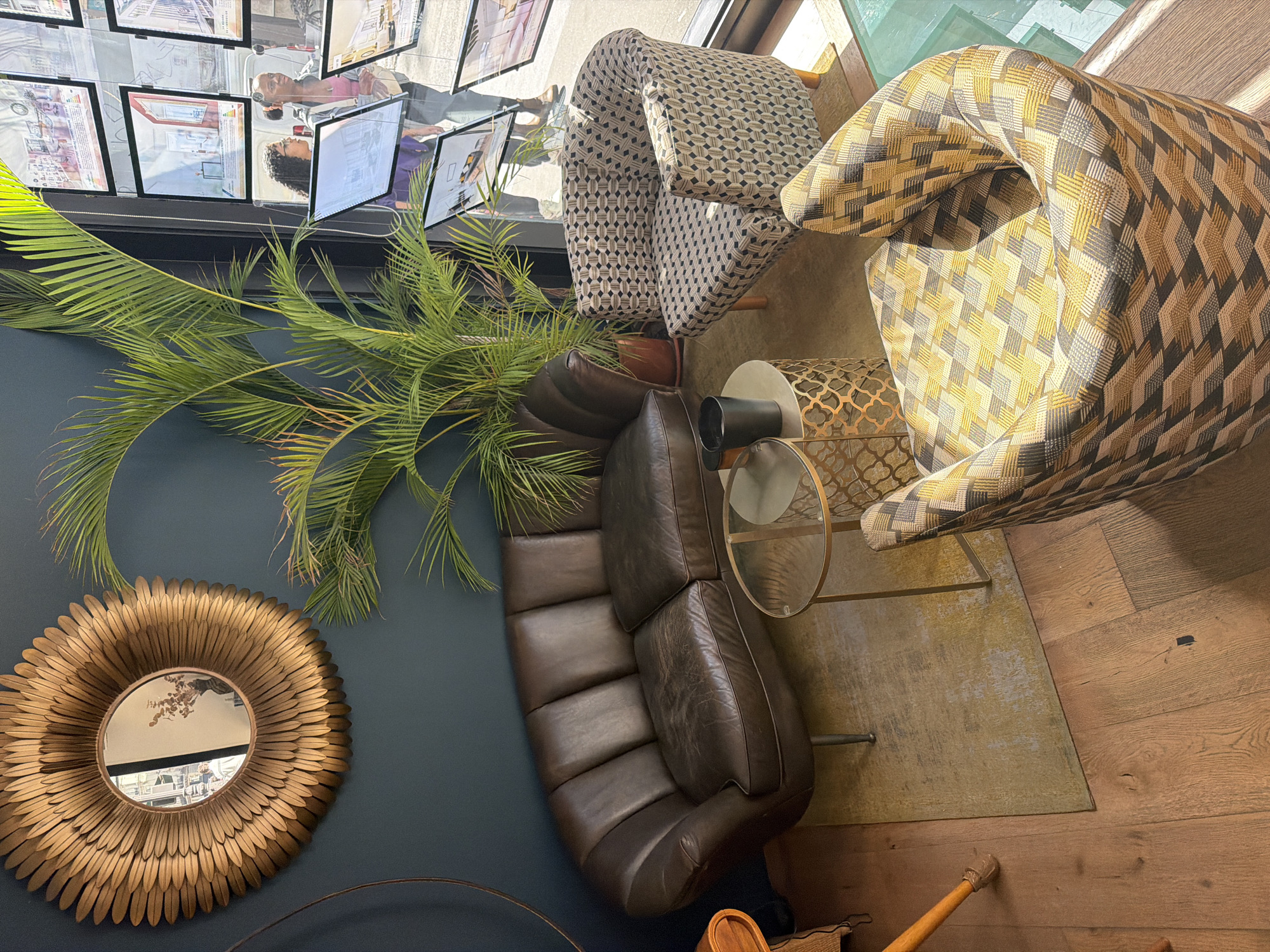 The scene is an interior shot with an oblique, high-angle perspective, likely taken from above looking down into the space. A deep blue wall is partially visible on the left, adorned with a round, ornate gold-colored mirror with radiating, petal-like embellishments. A tall, leafy potted plant, presumably a palm, extends its fronds both in front of the wall and towards the top of the frame.
In the center of the image, there is a dark brown leather sofa. The sofa is positioned adjacent to a modern glass and gold-colored side table. The table supports a black cylindrical object and is placed on top of a muted gold rug.
Two patterned armchairs, one in a geometric black and cream pattern and the other in a brown and gold, diamond-like design, are placed to the right and at the top of the frame. A hardwood floor extends beyond the rug.
At the top of the image, there is a display of framed posters or advertisements, possibly related to real estate, arranged in rows.
Given the real estate advertisements in the posters, the location is likely Paris, France.