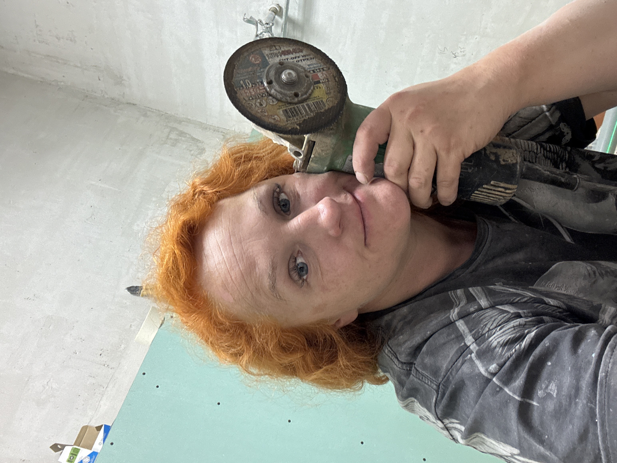 A person with reddish-orange curly hair is holding a handheld angle grinder close to their face. The person is wearing a gray patterned shirt and is positioned in what appears to be an interior setting with unfinished walls. A section of a light green or blue-green wallboard is also visible. The grinder appears to be well-used, with a visible abrasive cutting disc. It is not possible to verify the location of Chaikovsky, Russia based on the image alone.