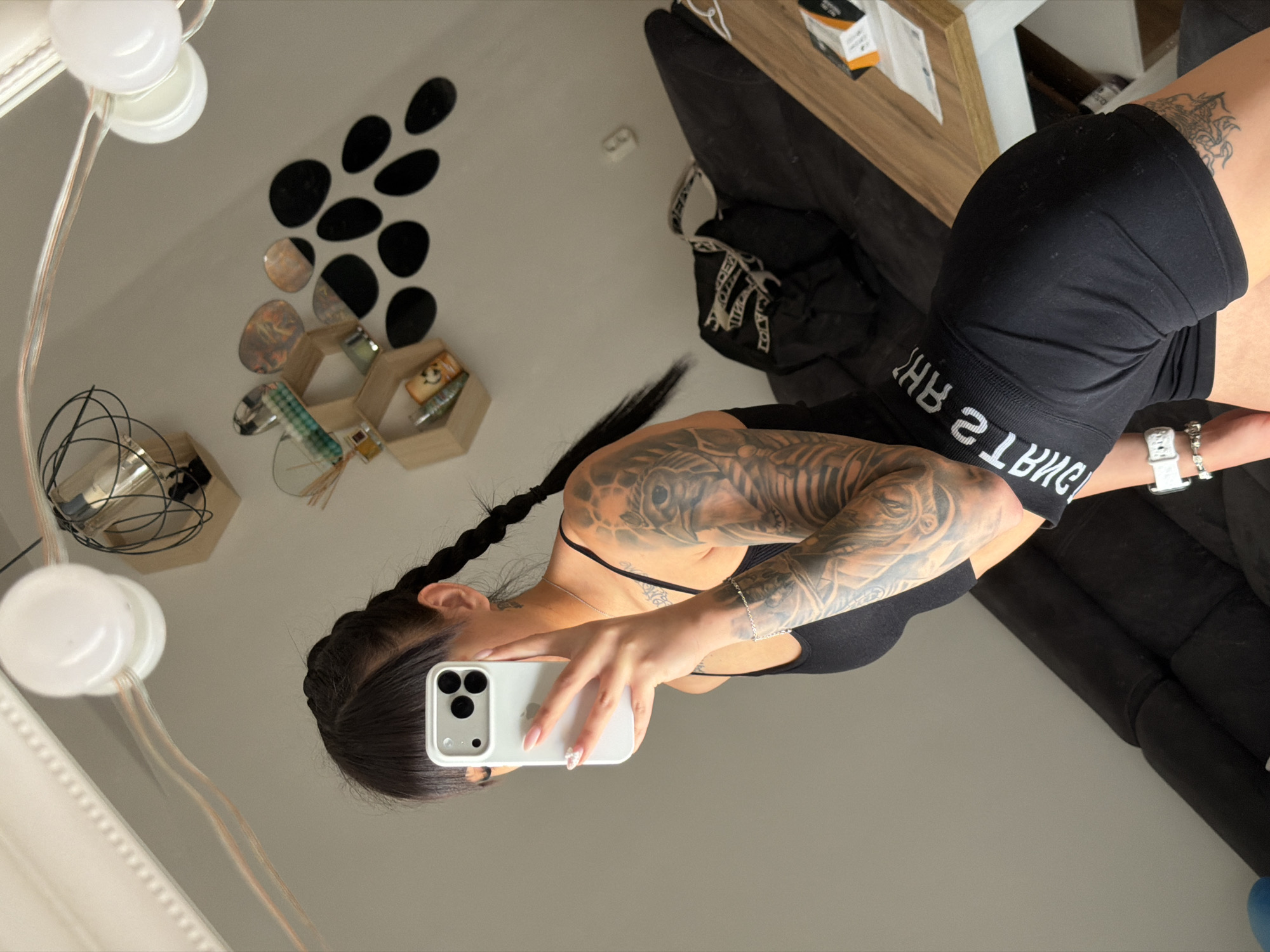 A young woman with dark hair in braids is taking a mirror selfie in what appears to be a bedroom. She is wearing a black sports bra and black shorts with a white waistband. Her left arm is covered in tattoos. She is holding a white iPhone in her right hand, obscuring her face. In the background, a black sofa is visible, along with a wooden table. The wall features decorative elements, including hexagonal shelves and round black decals. The lighting suggests an indoor setting, possibly a home. There is no indication of the location being in Plovdiv, Bulgaria.