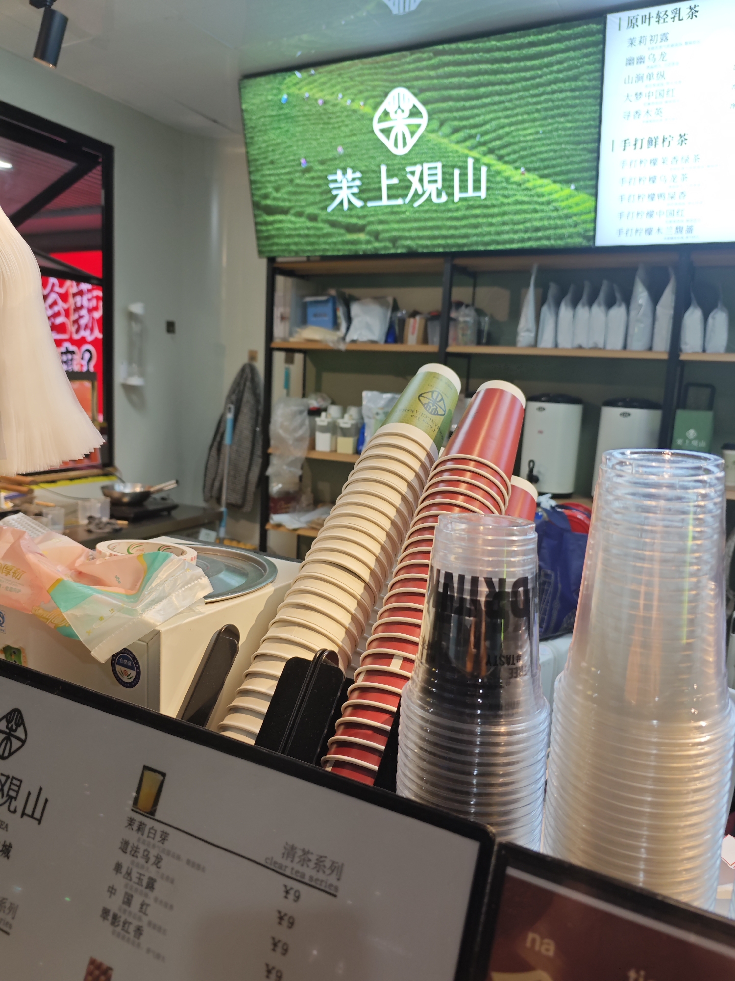 This is an indoor shot of a tea shop, likely in or related to the city of Liangjiang, China, based on the prominent signage in Chinese. The foreground features stacks of disposable cups in paper and plastic, some red and some clear, and a menu board with Chinese characters and prices. The menu lists various tea options, including "Original Leaf Light Milk Tea" and "Hand-Beaten Fresh Lemon Tea." In the background, there's a counter area with supplies, shelving with packaged goods, and tea-making equipment. A large LED screen displays a promotional image of a tea plantation and the name of the tea brand "茉上观山" (Mò shàng guān shān). The overall environment seems to be a tea shop or kiosk preparing beverages for customers.