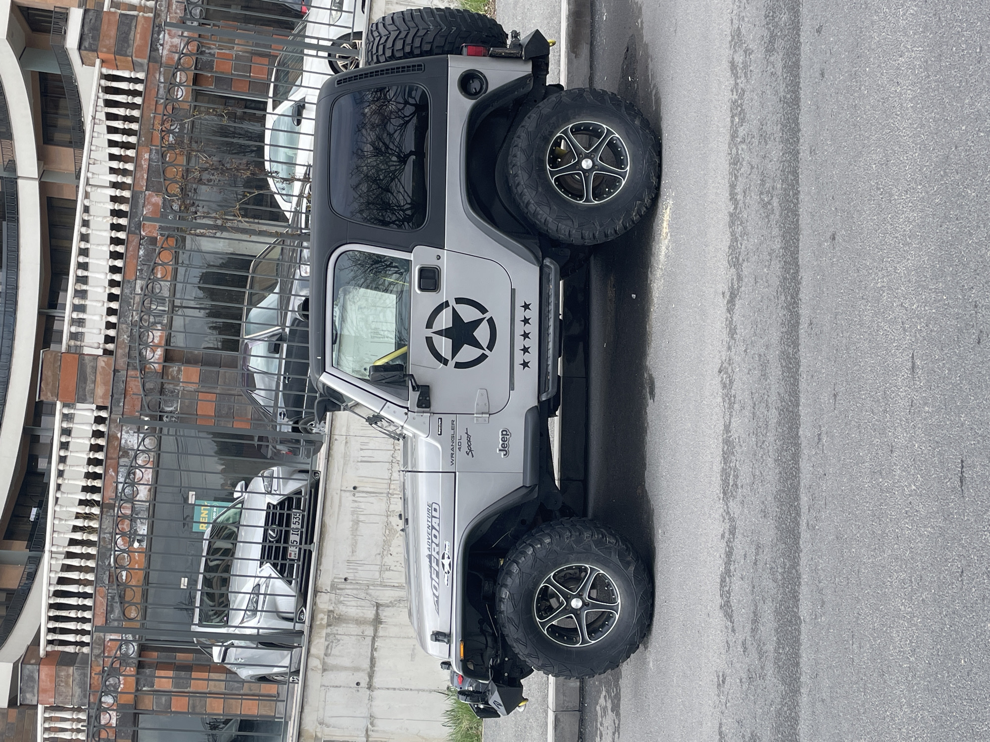 The image shows a silver Jeep Wrangler Sport parked on a gray paved street, likely in Yerevan, Armenia. The license plate of a silver Lexus SUV parked behind a black wrought-iron fence is visible: “35 IQ 535”. The Jeep has a black hardtop, a spare tire mounted on the back, and a large star decal on the driver's side door with five smaller star decals next to it. "ADVENTURE OFF ROAD," "WRANGLER 4.0L Sport," and "Jeep" decals are also visible on the vehicle's side. The scene appears to be in a residential or commercial area with a building visible in the background.