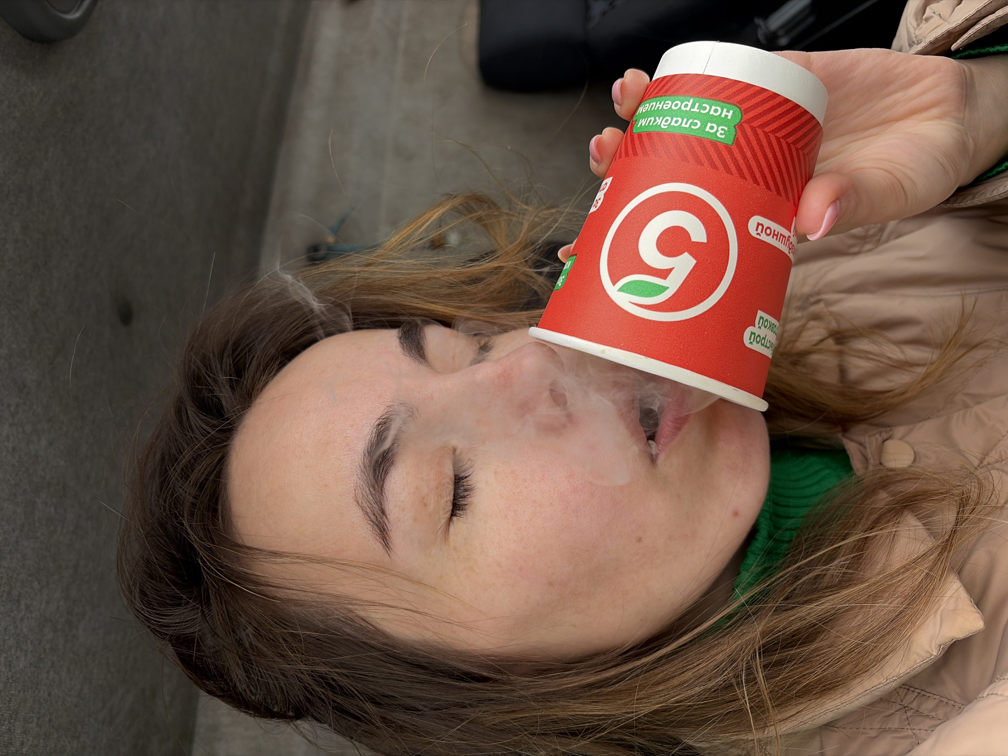 A woman is pictured in a sideways portrait. A paper cup with a red body and a white top is positioned over her mouth, and vapor is visible emanating from her lips. The cup is decorated with a large white circle enclosing a number "5" in red, with a green leaf detail. The text on the cup appears to be in Russian. The woman has fair skin, closed eyes, and light brown hair. She is wearing a green ribbed shirt under a tan quilted jacket. The background includes a gray surface, possibly the interior of a vehicle, and part of a black object. The presence of the "5" logo, common in Russian grocery stores, points to a probable location within Russia. The presence of Russian text on the cup further corroborates this hypothesis. The most probable location is within a car in the city of Izhevsk, Russia.