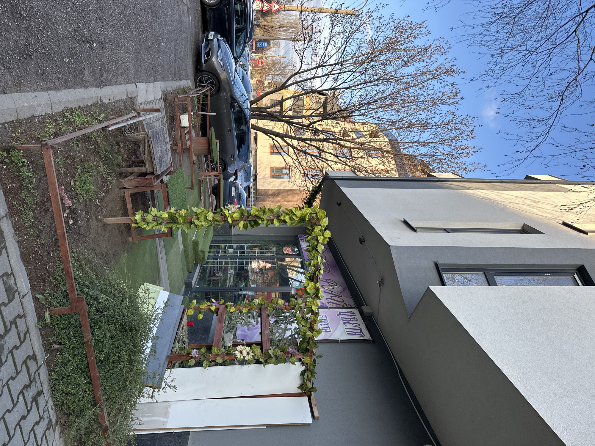 An exterior view shows the "Liberta" flower shop at an unspecified location in Sofia, Bulgaria. The building is modern with a gray facade. A sign with the shop's name is visible, adorned with artificial vines and flowers. A wooden structure, also decorated with artificial greenery, stands in front of the shop. The ground is a mixture of pavement, sparse vegetation, and artificial turf. A wooden bench and several wooden frame structures with no tops are to the left of the store. Cars are parked along the curb in front of the shop, and bare trees are visible in the background. The sky is blue and the lighting suggests it's daytime.