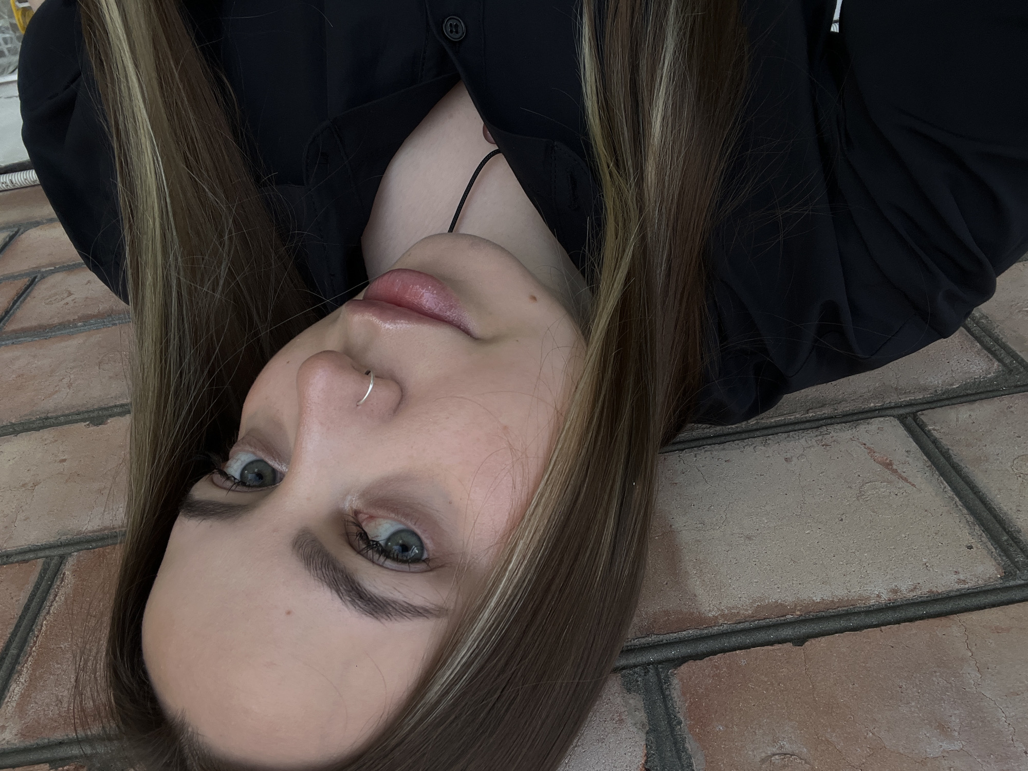 This close-up portrait captures a young woman with her head tilted to the right, possibly lying down or reclining, looking directly at the viewer. Her long, straight hair, which is predominantly brown with distinct blonde highlights, frames her face.
Her eyes are a striking green-blue, with visible eyelashes, and her eyebrows are well-defined. She has a silver hoop nose ring on her left nostril. Her lips appear full and are slightly parted, showcasing a natural or lightly glossed finish. She wears a black collared button-up shirt, which is partially unbuttoned, revealing a thin black string or necklace resting against her chest.
The background consists of brick paving or flooring with distinct grout lines, exhibiting shades of reddish-brown and gray. The lighting suggests an ambient environment, not harsh direct sunlight. No specific activity is clearly depicted other than posing for the photograph. The scene is likely set in Novopokrovskaya, Russia. There is no visible text in the image.