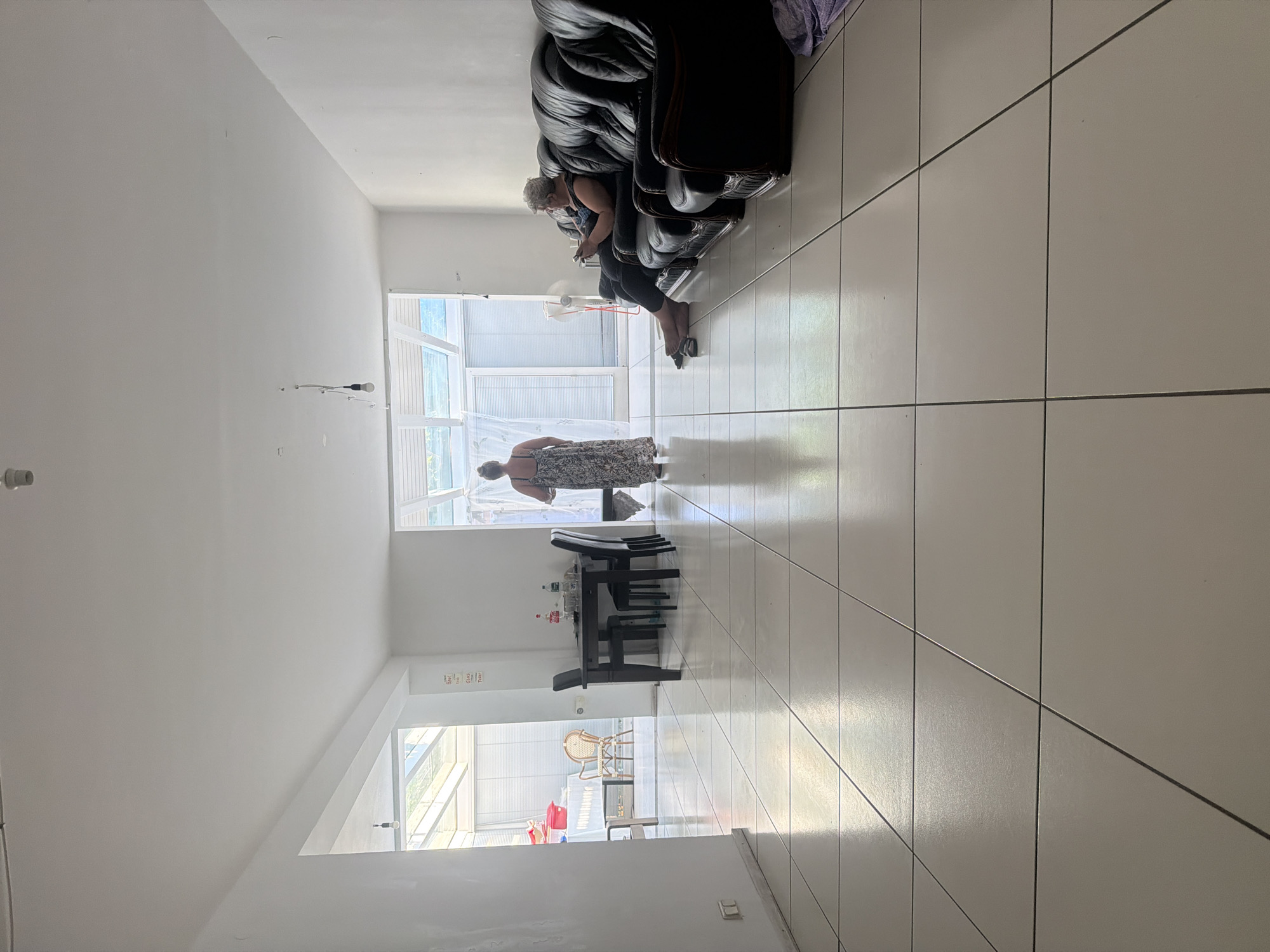 The image depicts a brightly lit, modern living space with large, reflective floor tiles. Two women are present. One woman, wearing a patterned dress, stands in the middle of the room, facing away from the camera and towards an open doorway. The other woman, with grey hair and wearing dark clothing, is seated on a black leather sofa on the right side of the room, looking down at a mobile device in her hands. A dark wooden dining table with four chairs is visible in the center left of the image. Beyond the dining table, through an open doorway, an outdoor area with a table and chairs, and a large red container are visible. The environment appears to be a residential interior, possibly a living room and dining area combined, with natural light streaming in from multiple openings, including a skylight-like structure above the doorway. There are no explicit visual cues within the image to suggest a specific location in Mannheim, Germany.