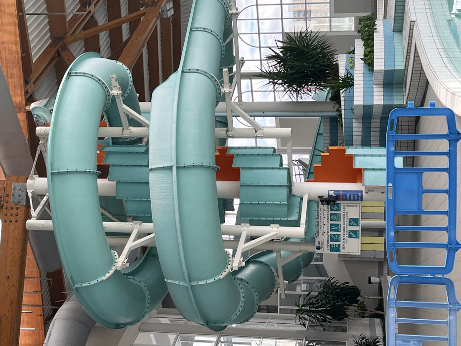 The scene is an indoor aquatic center located in Andrézieux-Bouthéon, France. A prominent teal-colored enclosed spiral water slide, constructed from segmented plastic sections, dominates the center of the frame, supported by white tubular columns and metal framework. An integrated spiral staircase, featuring orange treads and teal-colored risers, ascends alongside the slide. At the base of the structure, a white sign with French text titled "TUBOGGAN AQUATIQUE" and "INFORMATIONS GENERALES" displays safety pictograms, including a rule for '1 SEULE PERSONNE À LA FOIS' (1 person at a time). The facility features a high, pitched ceiling with exposed wooden trusses and white corrugated panels. Large arched windows along the right wall provide natural light and a view of distant urban buildings under a cloudy sky. The pool area in the foreground exhibits stepped edges with light blue and white striped ceramic tiles. Potted green plants are strategically placed around the base of the slide and within the tiled pool environment. A blue plastic safety barrier is positioned horizontally across the lower foreground, partially obscuring the pool. No individuals are present within the facility.