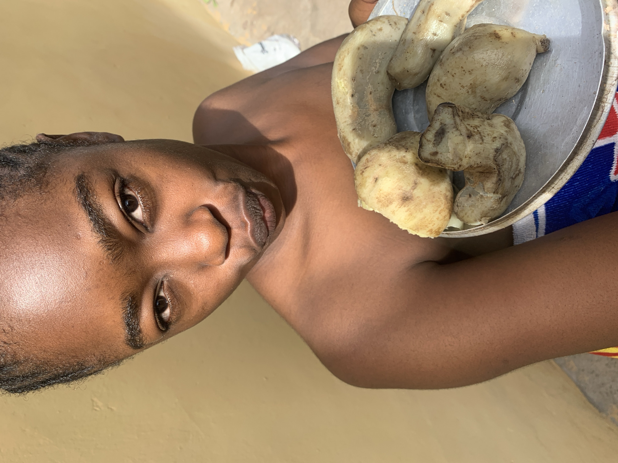 The image shows a close-up of a person's upper body, with a focus on their face and a metal plate filled with cooked food. The person appears to be of African descent, with dark skin and braided hair. They are looking towards the camera with a neutral expression. The food on the plate consists of several pieces of what appear to be boiled yams, with some dark spots on their skins.
The person is wearing a blue and red striped garment on their shoulder, partially visible under the plate. The background is out of focus, but it appears to be an outdoor setting with a light-colored wall and some sand or dirt on the ground. The lighting suggests it is daytime, possibly late morning or early afternoon, with harsh shadows indicating direct sunlight. The overall scene is rustic and appears to be in a rural or less developed area, consistent with the provided location context of Rafin Par, Nigeria. There is no visible text in the image.