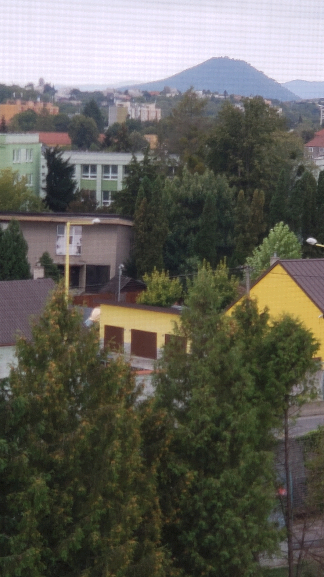 The image shows a view of a residential area in Prešov, Slovakia, with a mountain in the background. The foreground features several trees, including coniferous trees and deciduous trees. In the middle ground, there are various buildings, including a light green apartment building, a beige building with windows and a yellow pole, a yellow building with two garage doors, and a building with a dark brown roof. The background shows a hazy view of other buildings and a prominent mountain. The sky is overcast.