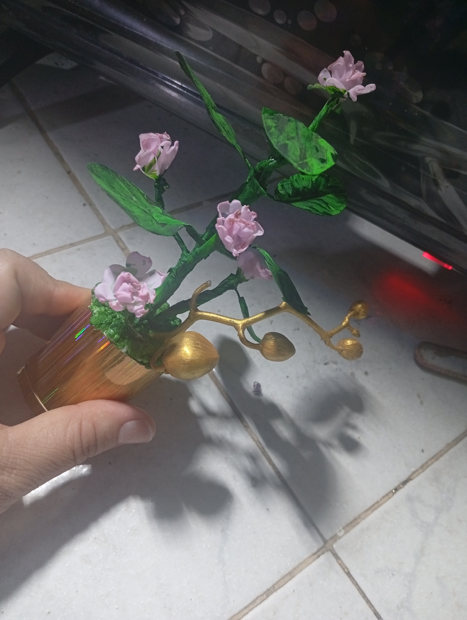 A close-up shot captures a person's hand holding a small, artificial floral arrangement. The arrangement features several pink flowers with green stems and leaves, supported by a gold-colored, cylindrical pot with a textured, iridescent surface. Green, moss-like material covers the base of the plant within the pot. A prominent golden, branched stem with multiple golden flower buds extends from the arrangement, adding a decorative element.
The hand and the object are positioned above a light-colored tiled floor with dark grout lines. In the background, a dark, reflective surface is visible, possibly furniture, with a bright red light emanating from its right side, suggesting an electronic device. Shadows of the hand and the plant are cast onto the tiled floor. The overall lighting appears dim, indicating an indoor setting, possibly during the evening or in an artificially lit environment. No discernible text is visible.