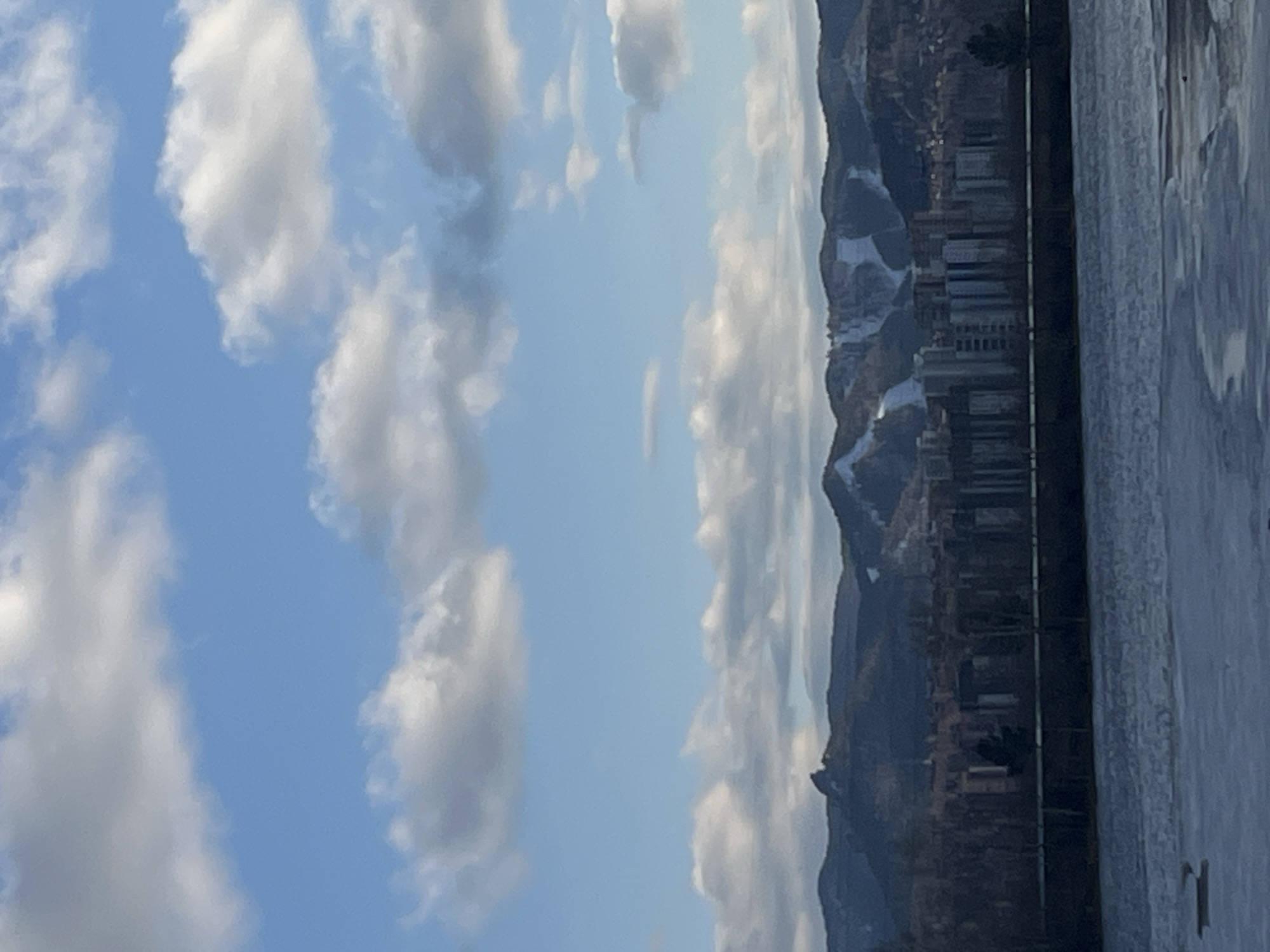 The image displays a panoramic winter or early spring landscape, presented in a portrait orientation that, when rotated 90 degrees clockwise, reveals a broad horizontal vista. The top two-thirds of the corrected image are dominated by a bright blue sky filled with scattered, fluffy white cumulus clouds.
Below the sky, a range of dark mountains or hills stretches across the mid-ground. These elevated landforms show distinct patches of white snow on their slopes and peaks. At the base of the mountains, an urban or semi-urban area is visible, characterized by numerous buildings, suggesting residential or commercial structures.
The foreground of the image features a large body of water, likely a river or lake, almost entirely covered in ice. The ice has a textured, uneven surface. A dark, relatively thin horizontal line runs across the frame, acting as a demarcation between the frozen water in the foreground and the distant cityscape and mountains. This line could be an embankment, a road, or a riverbank.
The scene is brightly illuminated, indicating daytime, likely mid-morning or early afternoon, under clear weather conditions with some cloud cover. No people, specific activities, or discernible text are visible. The overall atmosphere is one of serene vastness and a cold climate. Given the context, this is a landscape view of Krasnoyarsk, Russia, potentially showcasing the Yenisei River and its surrounding mountainous terrain.