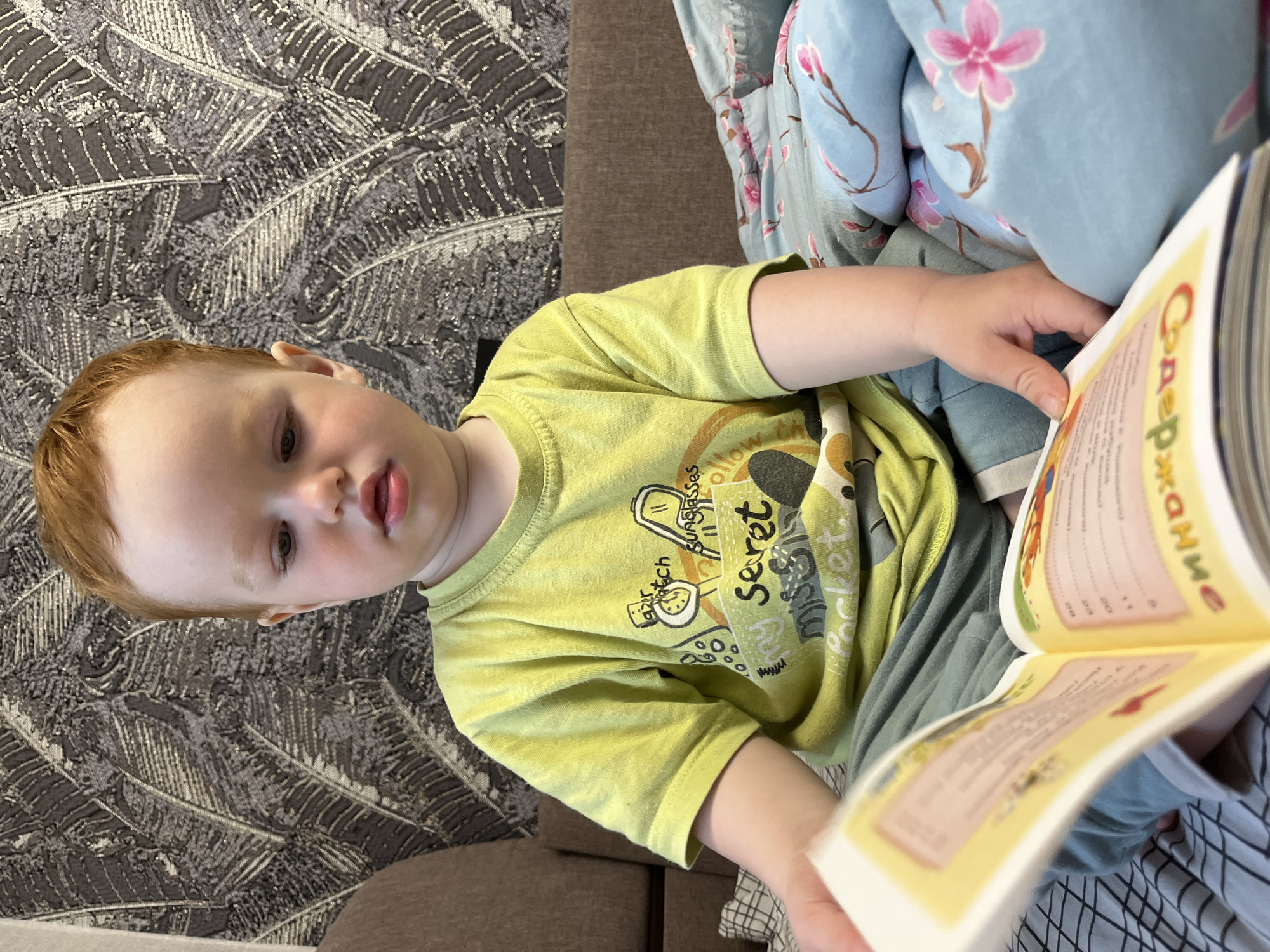 A young child with red hair sits on a bed with a book open in front of them. The child is wearing a light green t-shirt and blue pants. The background consists of a patterned wallpaper and a brown sofa. The bed has a blue comforter with a floral pattern. The book appears to be in Russian, with text visible on the pages. The scene is indoors, likely in a residential setting. There is no information available to determine the specific location in the city of Chaikovsky, Russia.