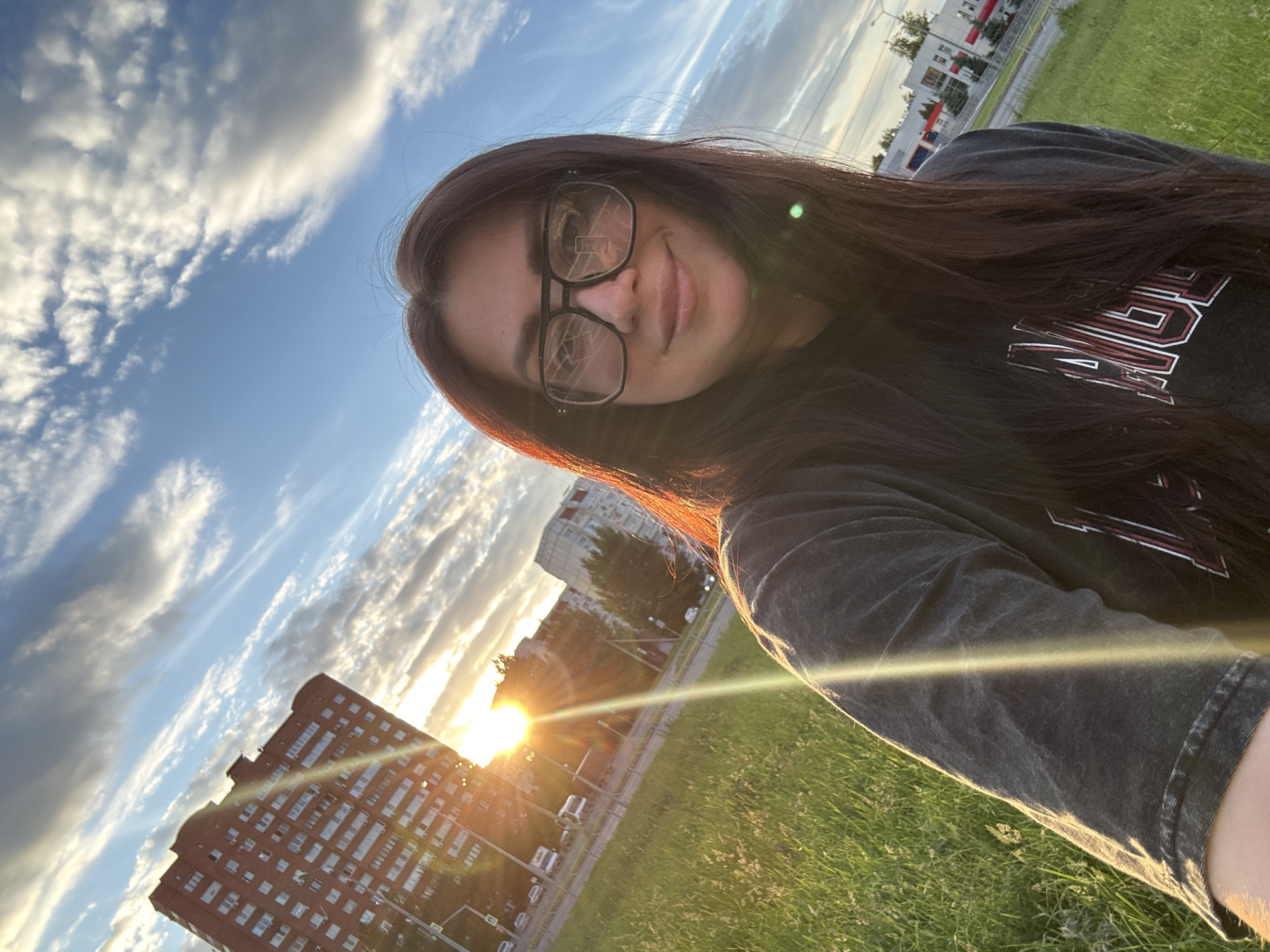 A woman with long dark hair and glasses is taking a selfie outdoors in what appears to be a city. She is wearing a dark grey t-shirt with lettering on the front. The background shows a tall brick building on the left, a street with cars, and a few smaller buildings and trees further in the distance. The sun is setting or rising, casting a bright glow and lens flare across the image, with a blue sky and wispy clouds above. The woman is smiling and looking towards the camera. The ground in the foreground is covered in green grass.