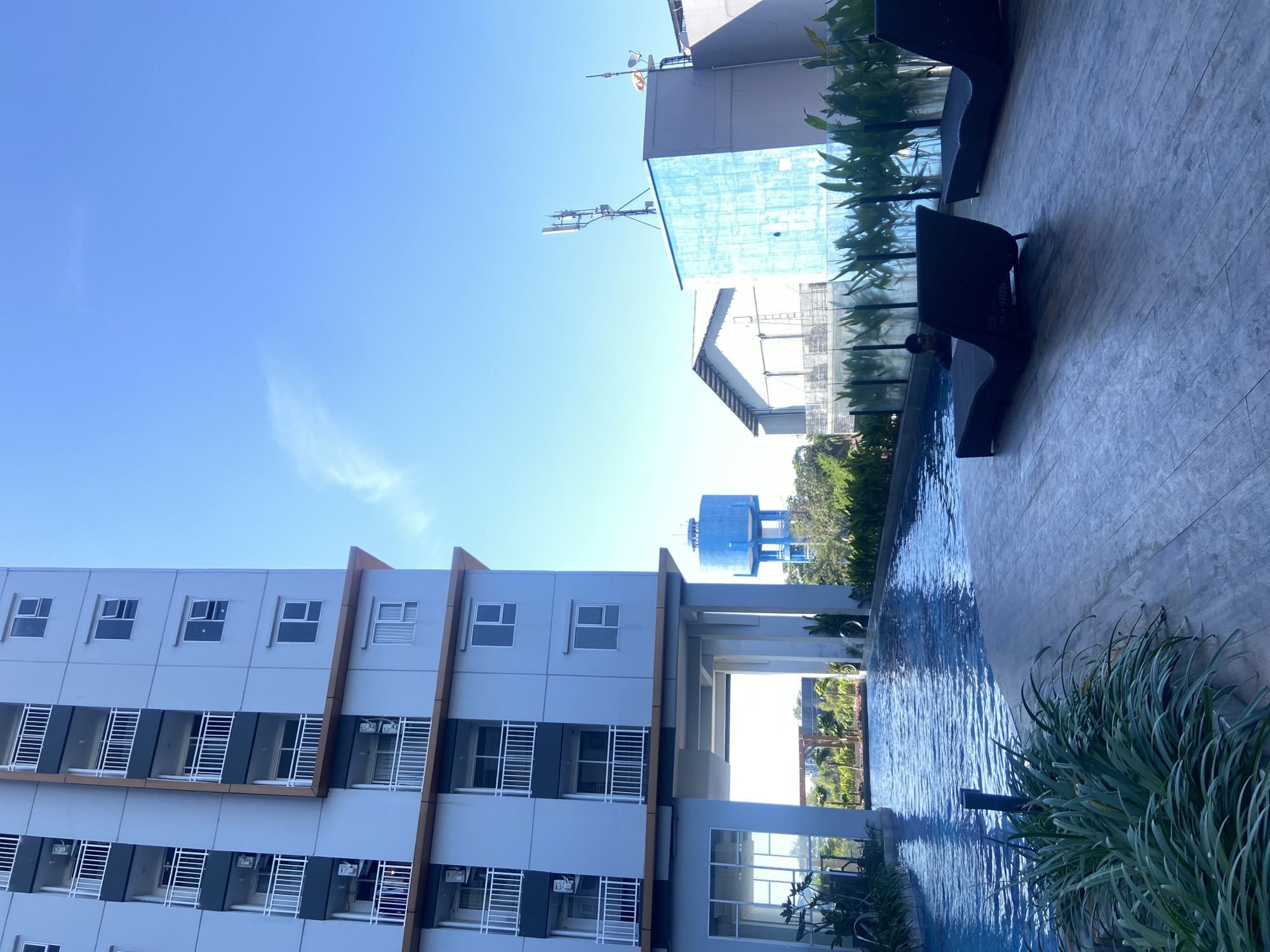 This image depicts an outdoor swimming pool area adjacent to a modern apartment building in Sidoarjo, Indonesia. The building, a tall, multi-story structure with a light gray facade and numerous balconies, dominates the left side of the frame. In the background, a blue water tower stands against a clear, bright blue sky, along with other buildings, including one with a light blue textured exterior and a white roof, and a cell tower. The pool itself is a prominent feature, with clear blue water reflecting the sky. Along the edge of the pool, on a wooden deck, are two dark-colored lounge chairs. A person is visible in the pool, and lush green foliage lines the pool and surrounds the deck. The scene appears to be bathed in natural sunlight, suggesting it is daytime.