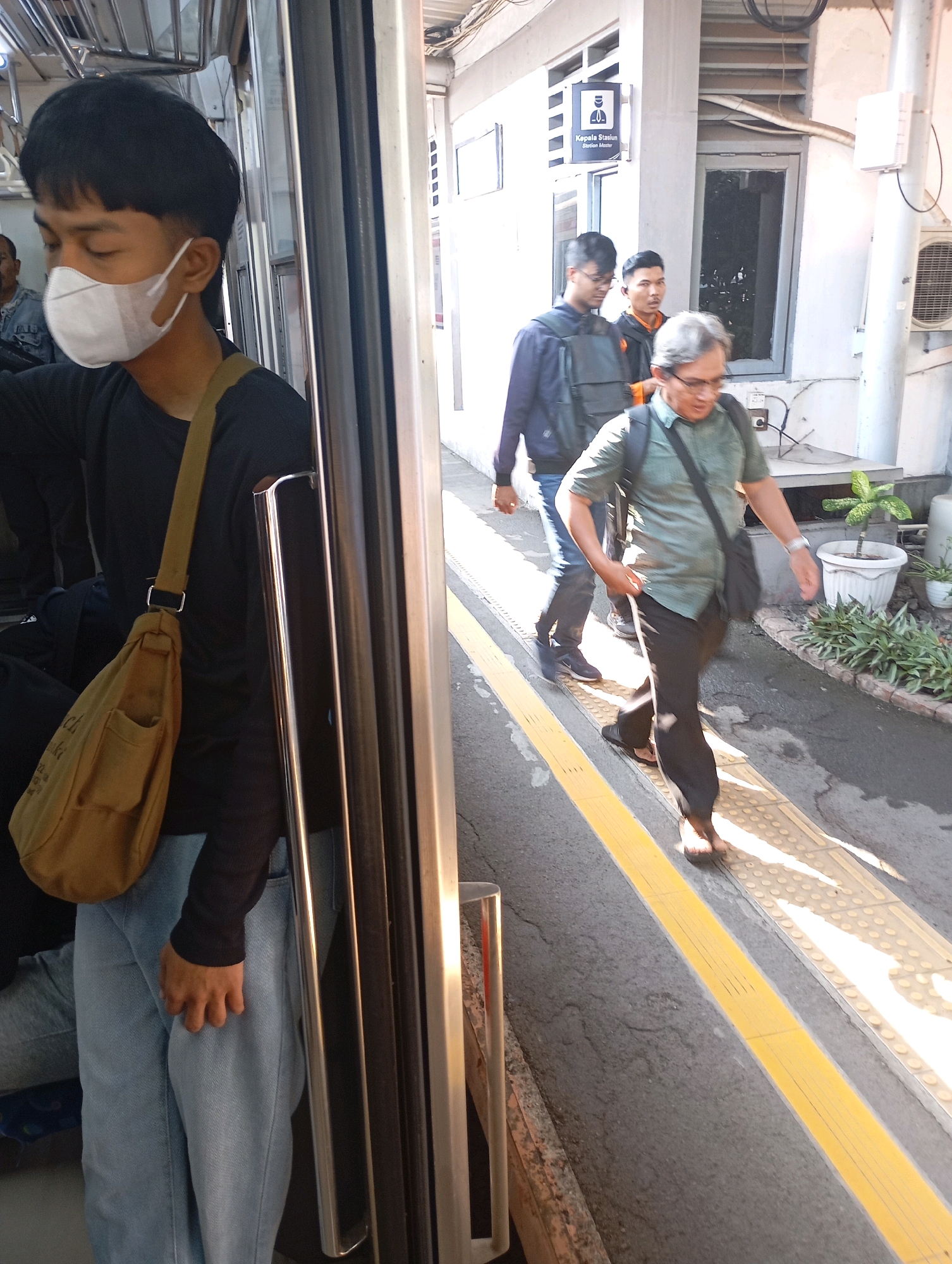 A young man wearing a white face mask and a black long-sleeved shirt is seen from the waist down, standing inside what appears to be a train carriage. He is wearing light blue jeans and a brown canvas shoulder bag. The train door is open, revealing a station platform with three other men walking away from the carriage. The man on the far right is older with graying hair, wearing a green collared shirt and dark pants, carrying a black backpack. Another man in a black jacket with a gray backpack is walking ahead of him, and the third man, wearing an orange and black jacket, follows. The platform has a yellow tactile paving strip and a white building with a sign that reads "Kepala Stasiun Station Master". The scene suggests a busy transit hub in West Jakarta, Indonesia, likely during the daytime, indicated by the natural lighting and shadows.
