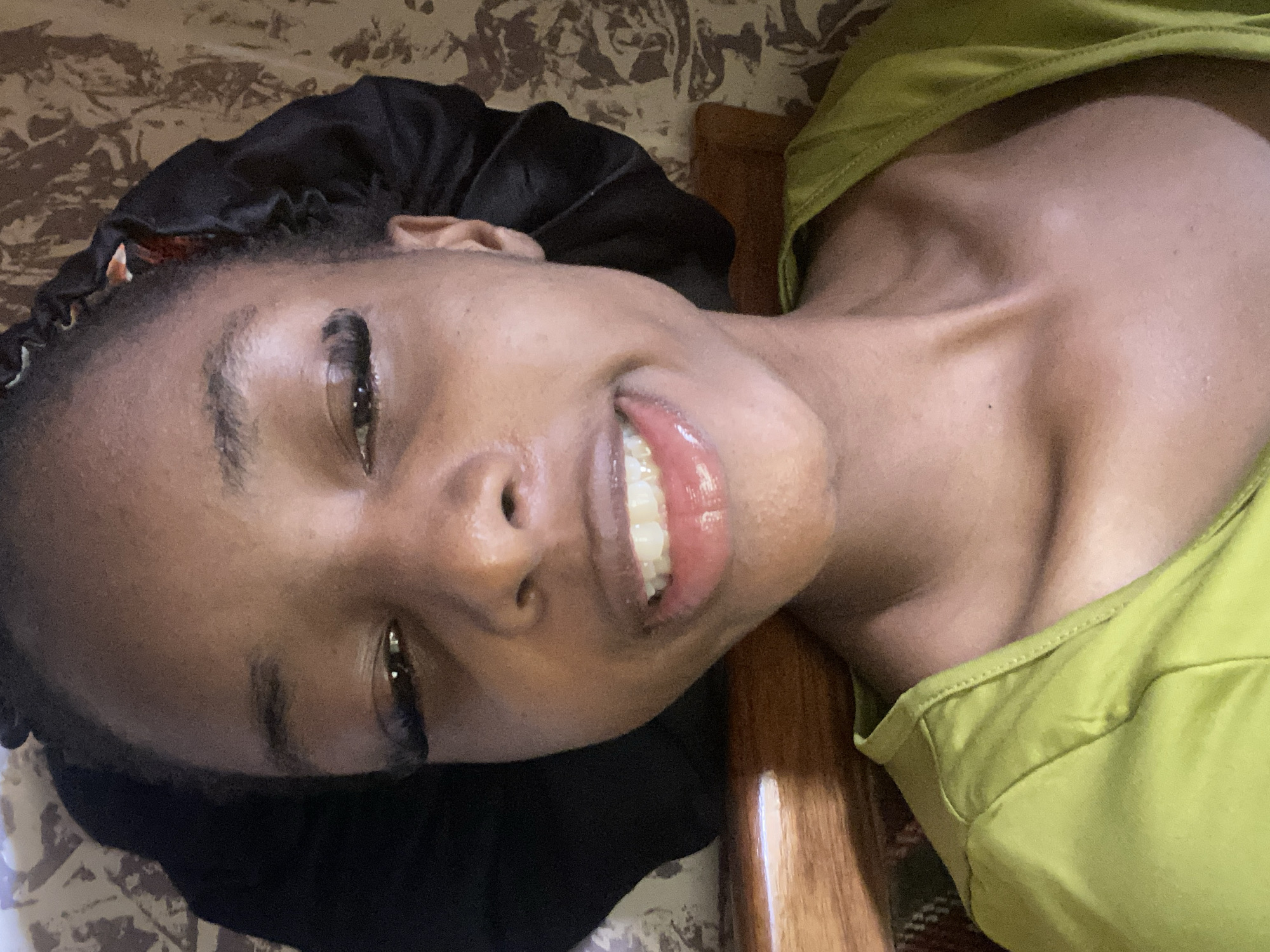 A close-up, rotated image shows a young Black person lying down, facing the camera. They are wearing a black satin bonnet on their head and a bright green sleeveless top. The person has a wide, genuine smile, revealing white teeth and glossy lips. Their eyes are open, looking directly at the camera, and they appear to be wearing false eyelashes. The background consists of a patterned fabric, possibly a bedsheet, and a portion of what looks like a wooden surface. The lighting suggests it is daytime. The overall mood is relaxed and cheerful. There is no visible text in the image. The location is identified as Jos, Nigeria.
