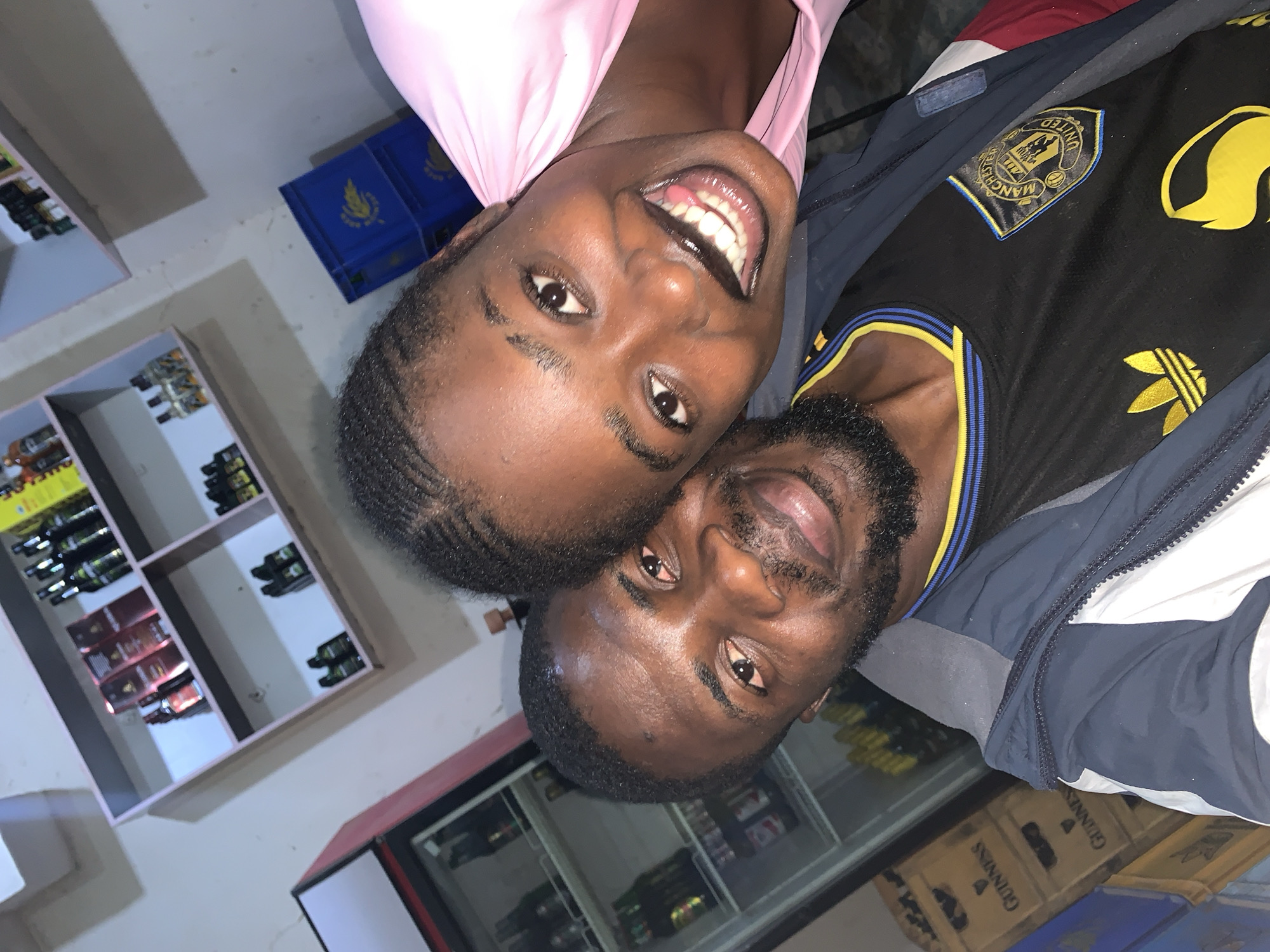 In New Stadium, Nigeria, a close-up selfie captures a Black couple. The man, with dark skin, a beard, and short hair, looks directly at the camera with a slight smile. The woman, also Black, with braided hair, smiles broadly, her teeth visible. She wears a pink top. The man is dressed in a Manchester United jersey and a jacket. Behind them, shelves are stocked with various bottles and boxes. A refrigerator is visible on the left, filled with drinks. Several stacked crates of Guinness are on the right, along with blue plastic crates. The overall setting appears to be a shop or convenience store.
