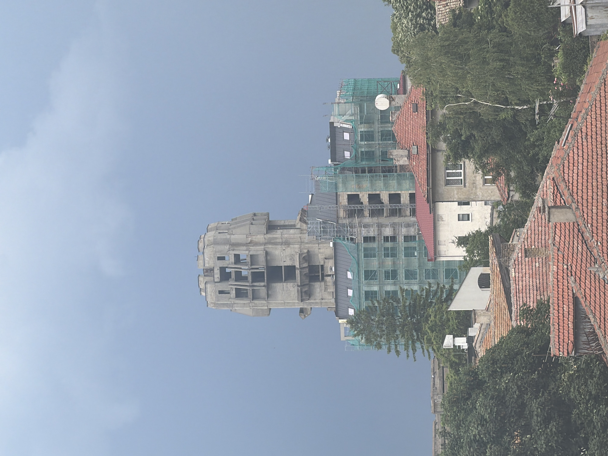 The image presents a panoramic, long-distance view of an urban landscape in Shumen, Bulgaria, under an overcast, light blue-grey sky.
Dominating the upper-mid portion of the scene is a large, unfinished brutalist concrete building, identifiable as the prominent landmark often referred to as the Shumen Hotel. This structure exhibits a distinctive, tiered architectural style with a broader, complex upper section featuring numerous exposed concrete walls and multiple empty, rectangular window openings, creating a somewhat fortress-like appearance. Its raw, unpainted concrete surface displays a uniform grey tone.
Below and to the sides of this central brutalist building, several other multi-story structures are visible. Many of these are partially covered by green construction netting and scaffolding, indicating active construction or renovation work. A tall, dark green coniferous tree stands in front of these mid-ground buildings, to the left of the brutalist edifice.
The lower half of the image is densely packed with residential buildings, characterized by their traditional, weathered red terracotta tiled roofs. Numerous chimneys, some with white caps, protrude from these roofs. Interspersed among these residences are mature deciduous trees with lush green foliage, partially obscuring the building facades. One particular house in the mid-ground features a light-colored stucco facade and white-framed windows. A white, circular satellite dish is mounted on a building roof in the mid-ground, to the right of the central axis.
The sky, occupying the upper third of the frame, is a consistent, muted blue-grey, suggesting heavily overcast conditions with no direct sunlight. No human figures, vehicles, or specific activities are discernible within the frame. The overall scene conveys a mix of established residential areas, ongoing urban development, and a significant, imposing brutalist structure, all under a subdued ambient light.