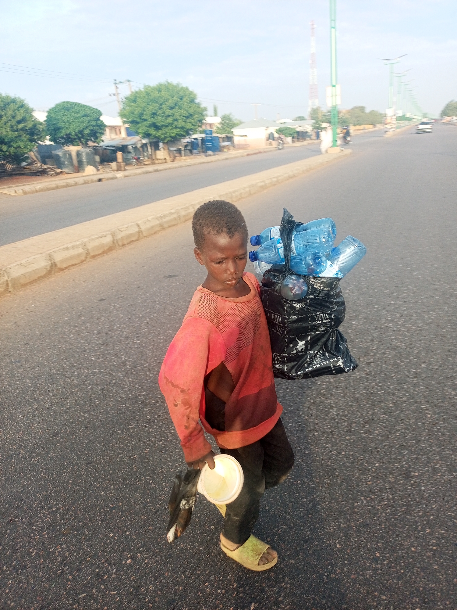 A young boy walks on a paved road with a bag of plastic water bottles slung over his shoulder, and a white bowl in his left hand. He is wearing a faded red, long-sleeved shirt that is torn at the arm, dark pants, and green sandals. The road has a cement shoulder along the roadside. Beyond the road is a row of trees and single-story buildings. In the distance, a tall communications tower is visible, as well as street lights and sparse vehicular traffic. The boy is in Tumfure, Nigeria.