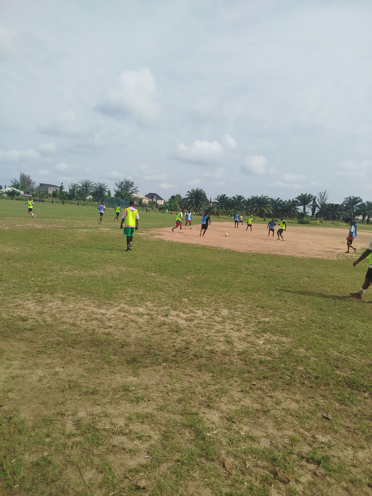 The image shows a soccer match in Ikot Mbonde, Nigeria. The location is a large, grassy field with a visible dirt patch in the center. Several people are playing soccer, wearing various colored shirts and shorts. Buildings and trees are visible in the background, under a cloudy sky.
