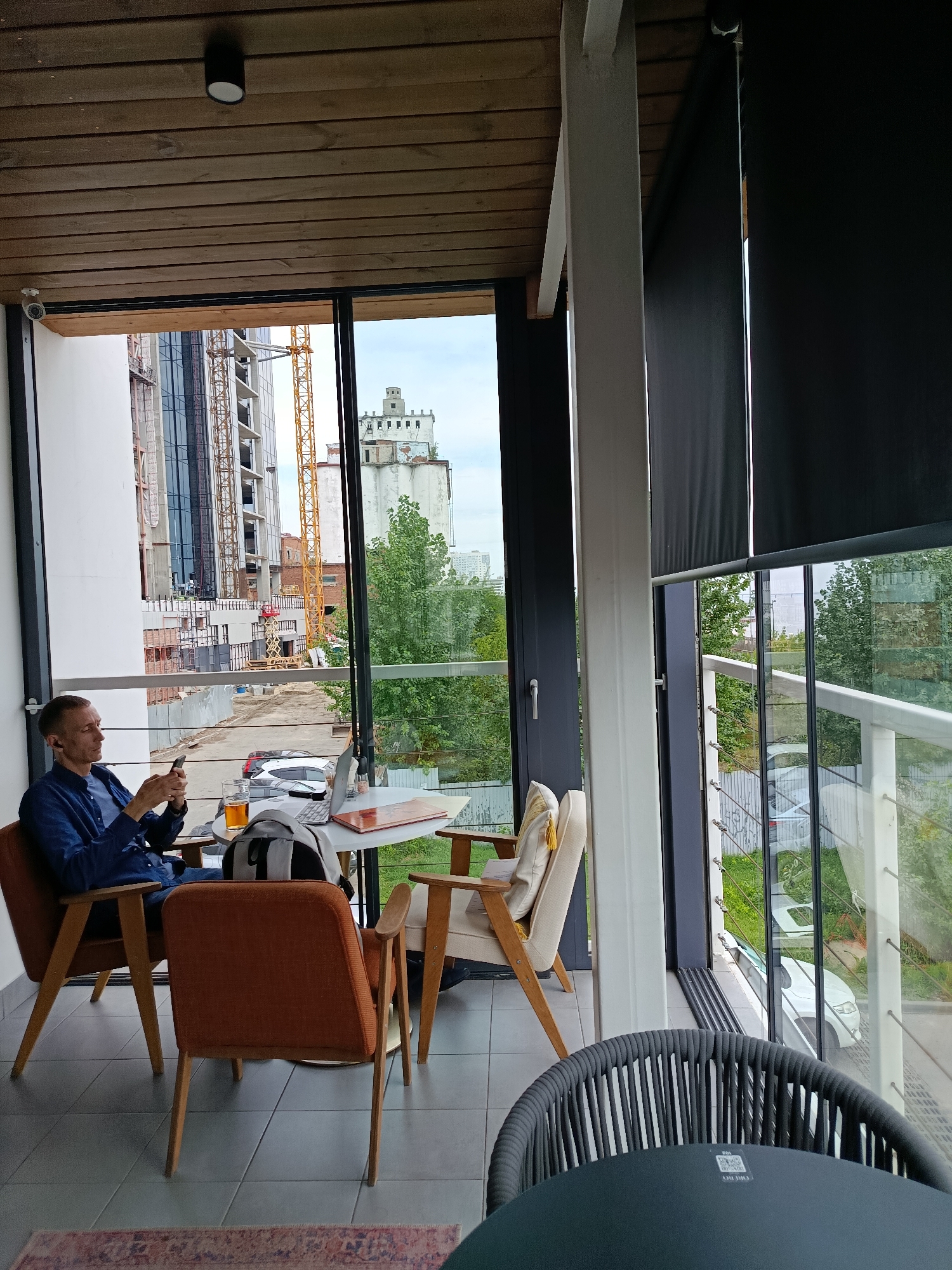 The image captures a man sitting at a table on a balcony overlooking a construction site in Saratov, Russia. The man, wearing a blue shirt, is looking at his phone and holding a glass of amber liquid. On the table in front of him are a laptop, papers, and a backpack. The balcony is furnished with chairs and a table, and a dark-colored outdoor chair and table are in the foreground. Beyond the balcony, a multi-story building is under construction, with cranes and scaffolding visible. The sky is overcast. The architectural style of the building in the background, an old concrete structure with a distinctive tower, suggests it may be a repurposed industrial building.