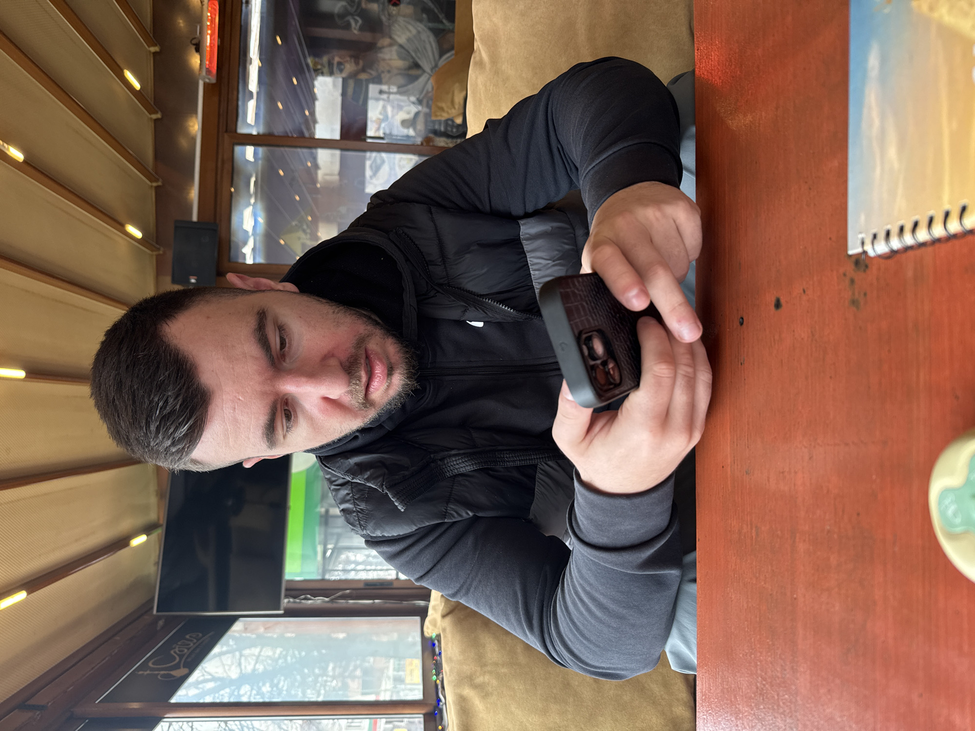A man with short dark hair, a beard, and wearing a black vest over a long-sleeved gray shirt is sitting at a wooden table and looking at a cell phone in his hands. The table is brown and has a spiral-bound notebook on it. The man is sitting on a tan cushioned bench. Behind him is a window with horizontal blinds and a TV on the wall. The picture was probably taken in a cafe or restaurant, potentially in Sofia, Bulgaria as suggested by the location tag. The precise location within Sofia is unknown without further information.