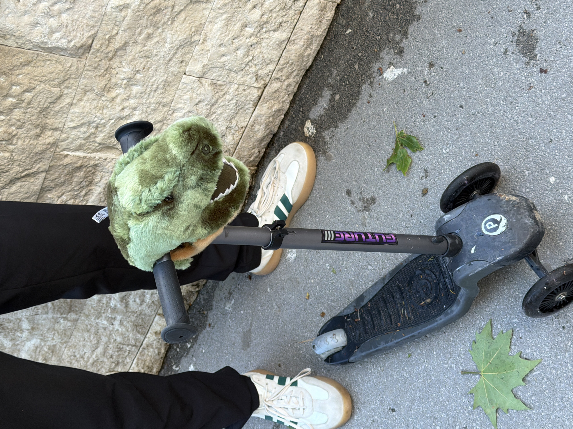 The image depicts a person standing on a sidewalk next to a stone wall in an urban environment, possibly Budva, Montenegro, given the context. The person is wearing black pants and white sneakers with green stripes. They are holding the handlebars of a gray children's scooter with a green stuffed dinosaur head attached to one of the handlebars. Two green leaves are scattered on the gray sidewalk near the scooter.