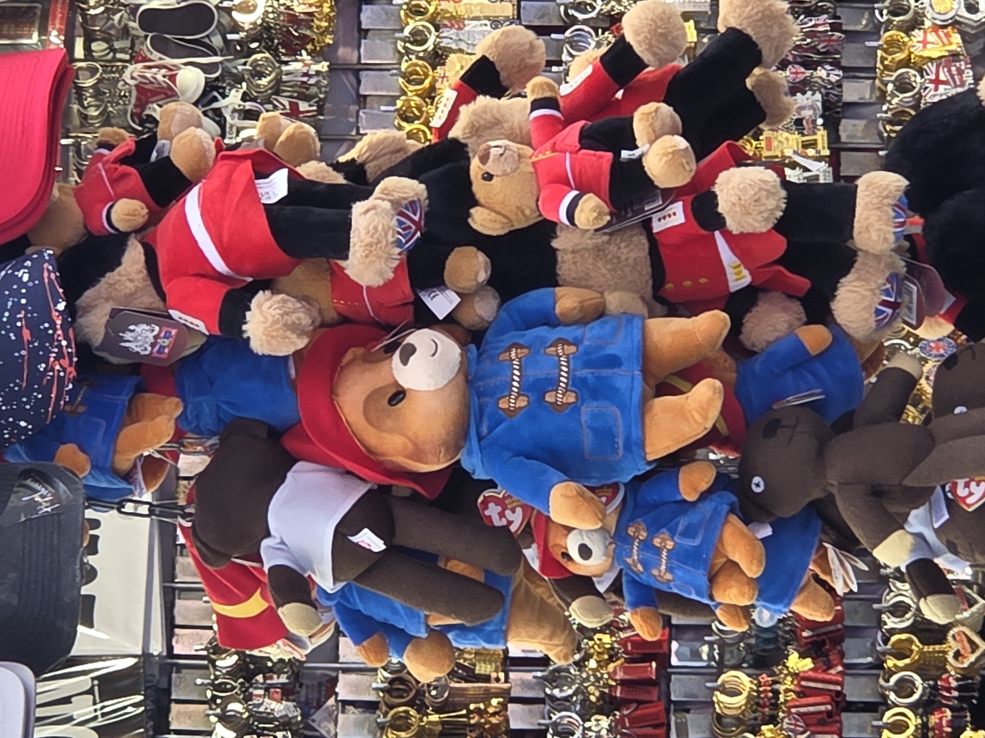 The image shows a display of teddy bears and souvenirs in a shop, likely in London, United Kingdom.
Several Paddington Bear plush toys, some wearing blue duffel coats, are centrally located. Other bears are dressed as guards in red jackets and black trousers. These toys hang from metal hooks, suspended in front of a wall with other souvenirs, including keychains featuring Union Jack flags, London symbols, and other tourist merchandise. The background is filled with these items, suggesting a souvenir shop catering to tourists.