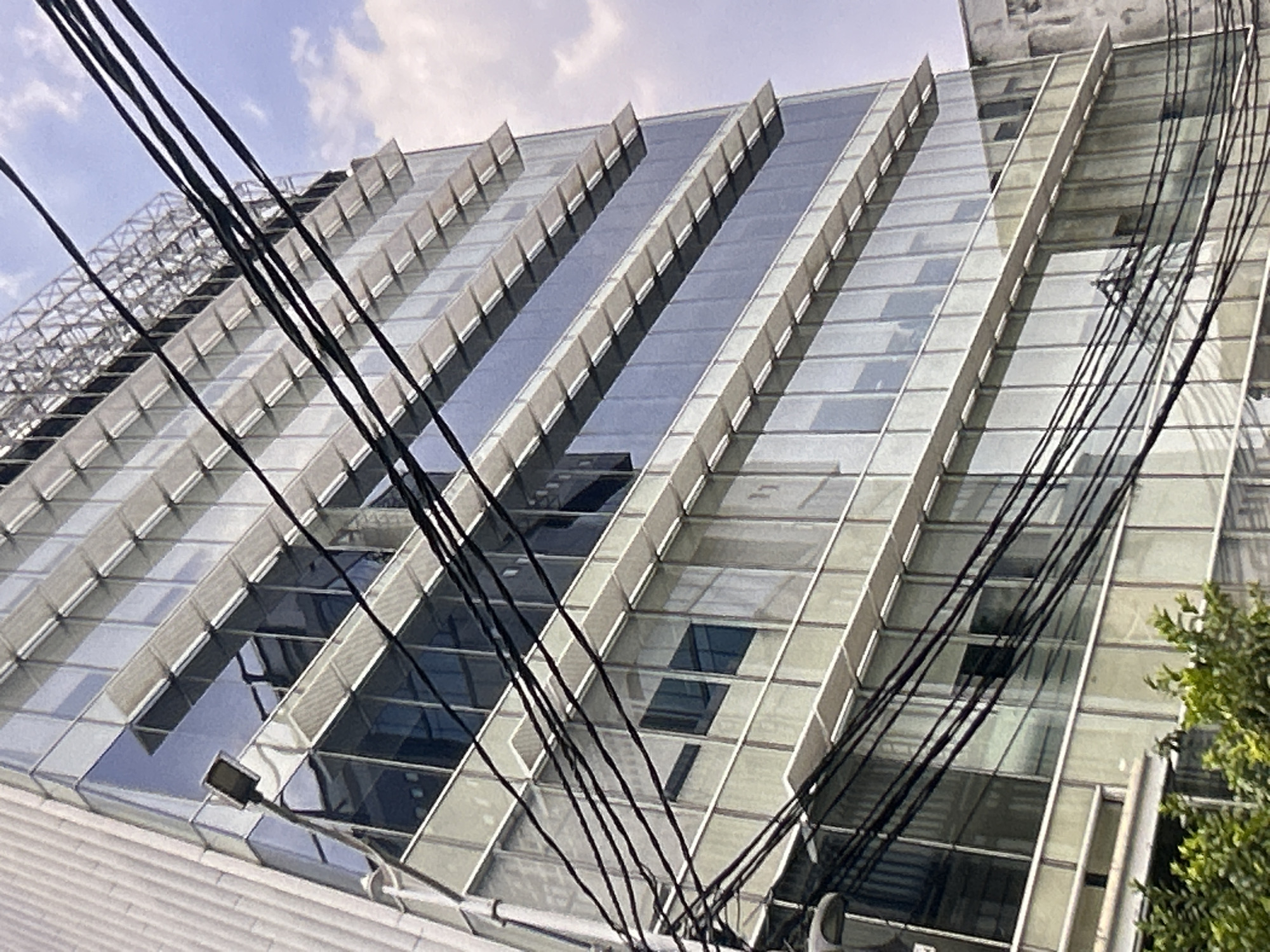 The image shows a modern, multi-story building with a glass facade and horizontal sunshades on each floor. The building is tall and appears to be located in a city. Several thick black wires are visible in the foreground, crossing diagonally across the frame and obscuring parts of the building. The sky above is cloudy. The image is shot from a low angle, looking up at the building. There are no people or vehicles clearly visible in the image. The building's reflective windows show glimpses of the sky and interior structures, but no clear details of occupants or activities. The overall impression is one of urban architecture, with a focus on the building's design and the foreground obstruction of the wires.