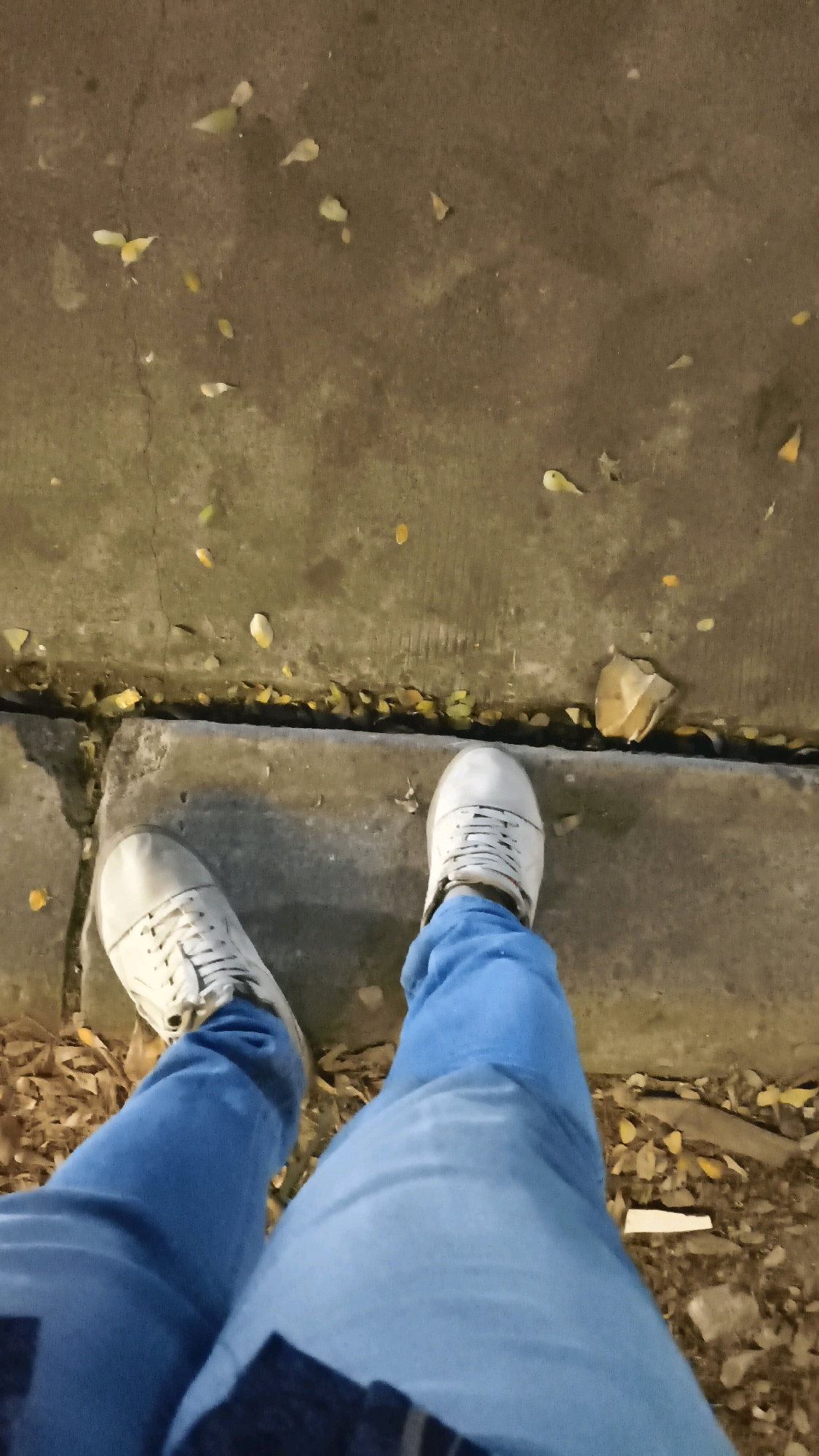 The image shows a first-person perspective of someone standing on a sidewalk in Karangsatria, Indonesia, at night. The person is wearing light-colored sneakers and blue jeans. The sidewalk is made of concrete slabs, with a curb separating it from a dirt area covered in fallen leaves and a crumpled piece of paper. The lighting suggests it is an outdoor urban environment.