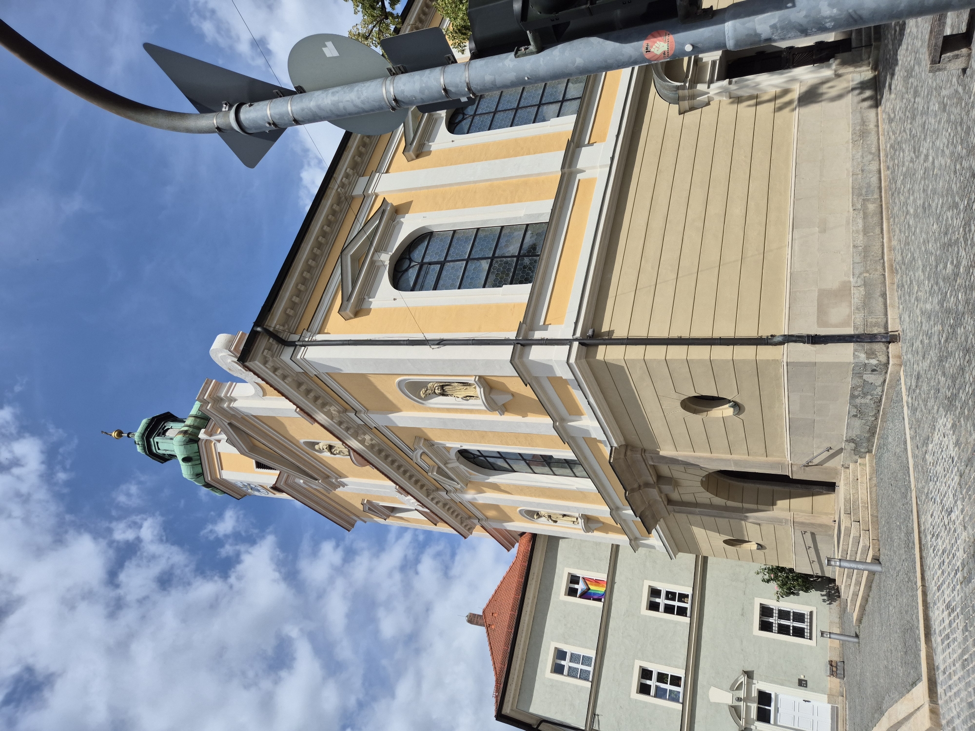 This is a medium-angle, exterior, ground-level shot of the St. Georgenkirche in Regensburg, Germany, under a partly cloudy sky. The church, painted yellow with white accents, features a green domed tower with a clock. A rainbow flag is visible in the window of a light gray building to the left of the church. In the foreground, a cobblestone street and sidewalk lead up to the church's entrance, which has a stone staircase and arched doorway. A metal traffic light pole with multiple signs stands to the right of the frame, partially obscuring the church's facade.