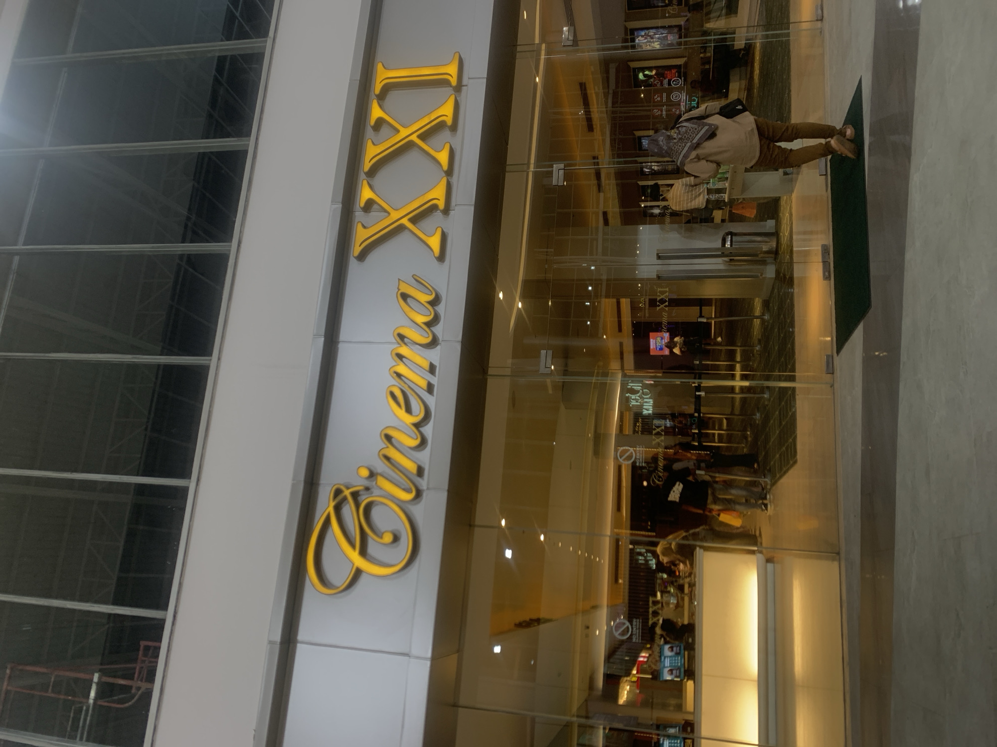 This image captures the entrance of a "Cinema XXI" movie theater, likely located within a modern shopping complex in Samarinda, Indonesia. The dominant feature is the large, three-dimensional sign with the name "Cinema XXI" displayed in gold-colored lettering on a grey facade. The word "Cinema" is in a flowing script font, while "XXI" is in a bold serif style.
In front of the cinema entrance are clear glass doors, reflecting the interior lighting and a partial view of what lies beyond. A person, wearing a light-colored top, brown trousers, and a patterned headscarf (hijab) with a dark shoulder bag, stands facing these doors, positioned on a dark green doormat. Their posture suggests they are either entering or waiting to enter the cinema.
Through the glass, reflections show parts of the cinema lobby, including glowing screens that appear to display movie information or advertisements. Other faint reflections of people and interior structures are also visible. On the glass doors, the text "Cinema XXI" is visible in reverse due to reflection, alongside a "no entry" or "no specific item" symbol. To the left of the cinema entrance, a large windowed section of the building reveals internal support structures and what appears to be some scaffolding. The scene is brightly lit by artificial indoor lighting, giving no clear indication of the time of day beyond being an active commercial environment.